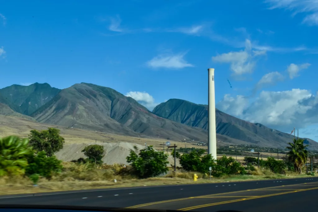 Une grande cheminée blanche se dresse près d'une route avec des lignes de séparation jaunes, entourée d'une végétation clairsemée et de palmiers avec des montagnes vertes et un ciel bleu en toile de fond.