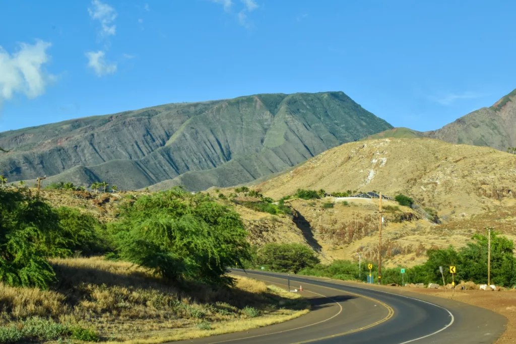 Une route pavée serpente à travers un paysage sec avec des buissons verts dispersés menant vers de grandes montagnes escarpées sous un ciel bleu clair.