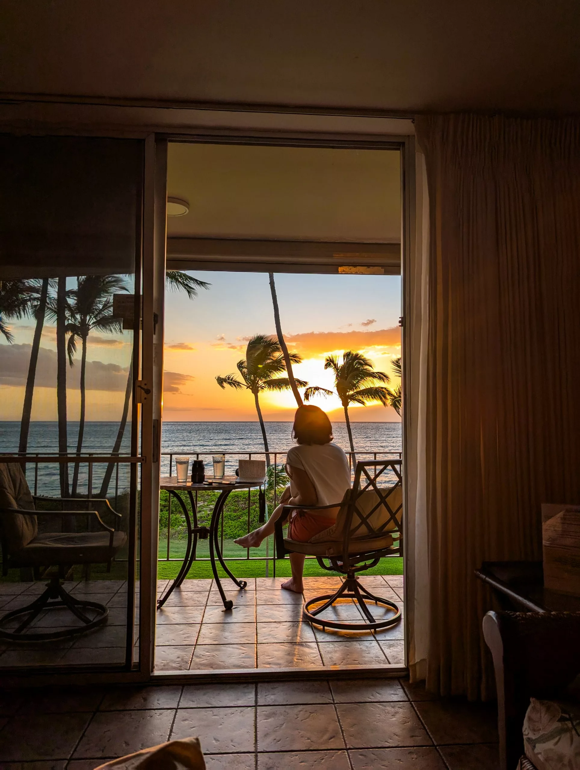 Une personne est assise sur une chaise de balcon face à l'océan au coucher du soleil, avec des palmiers