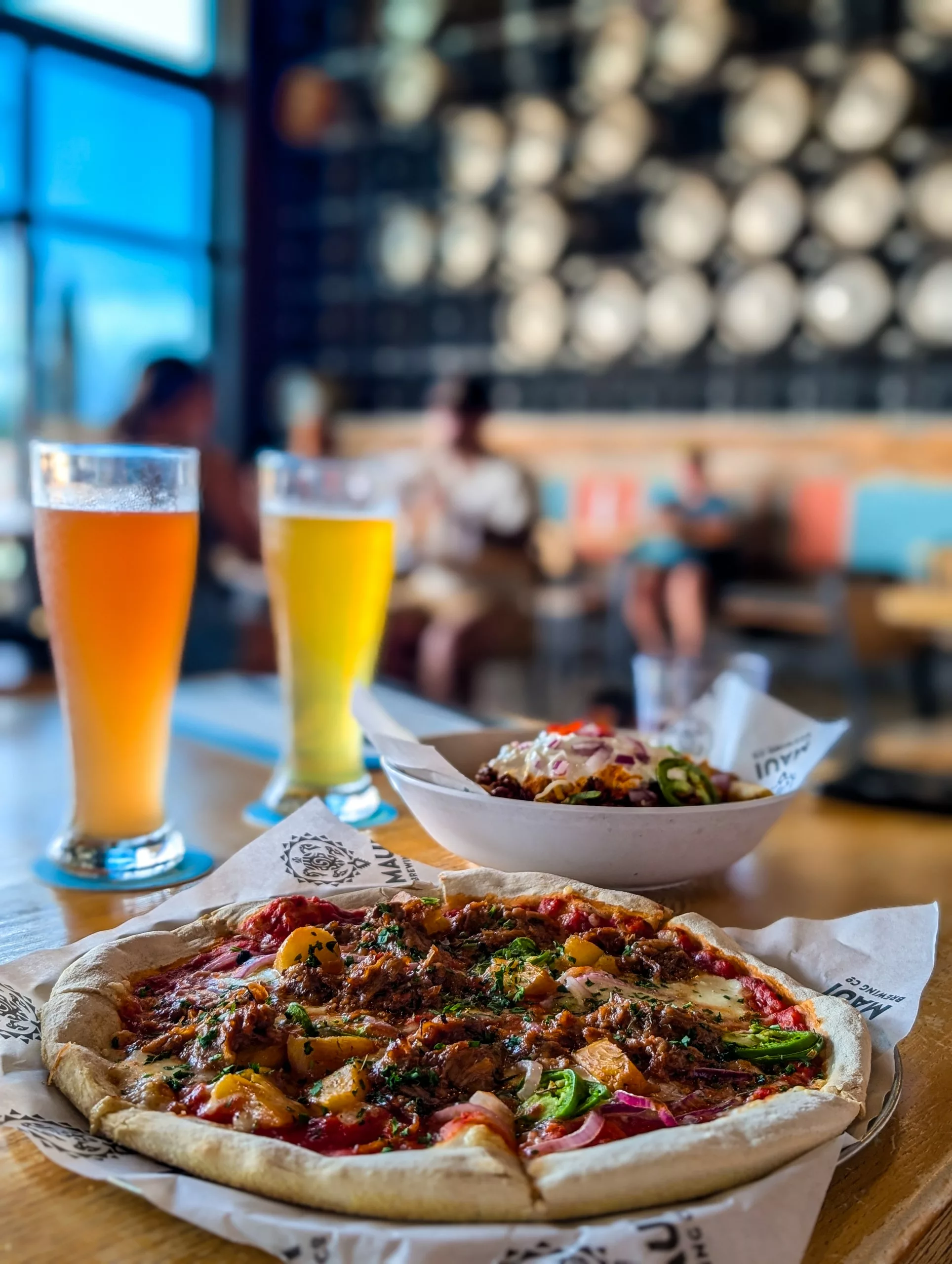 Une pizza garnie d'herbes et de légumes est posée à côté de deux grandes bières, dans un restaurant moderne avec des gens flous en arrière-plan.