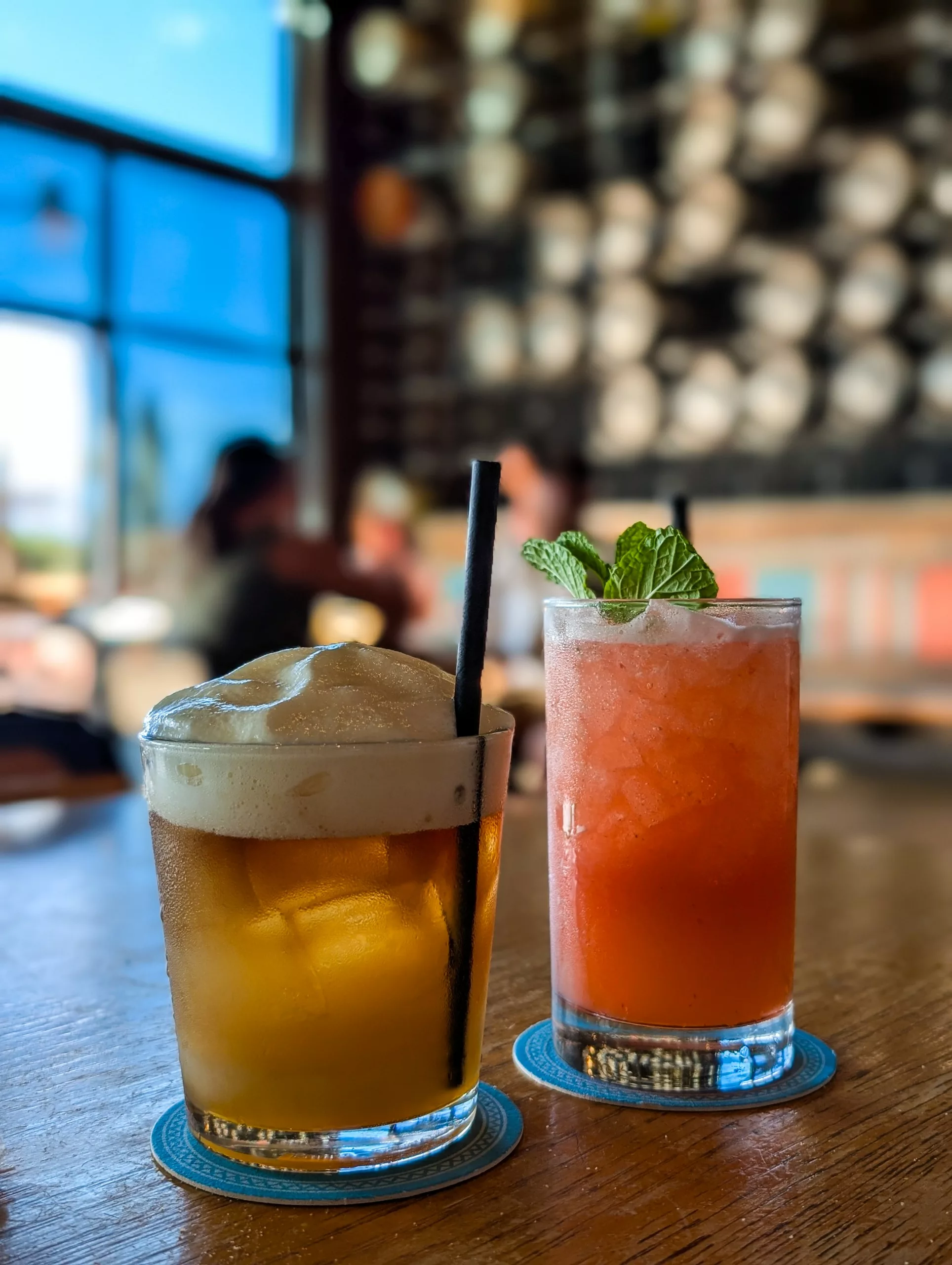 Deux boissons sur une table en bois : l'une est une boisson mousseuse brun clair, l'autre une boisson glacée rose-orange à la menthe, sur fond flou de personnes et de décor.