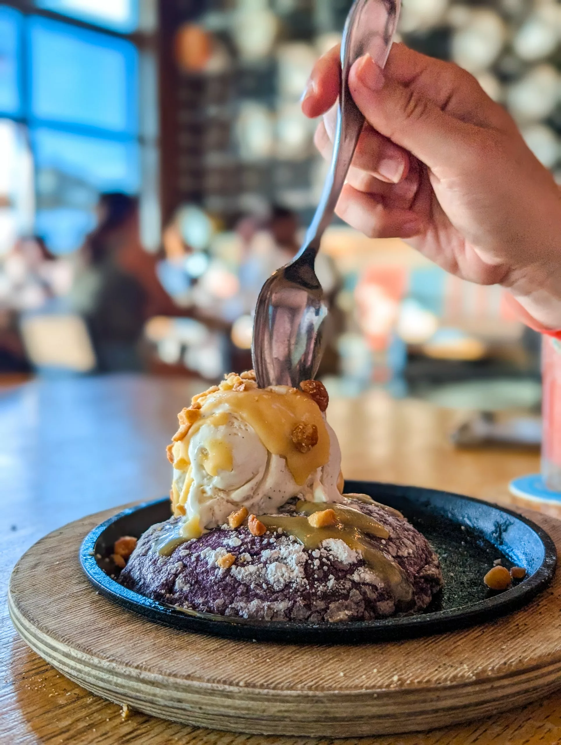 Une main tient une cuillère sur un dessert avec de la glace à la vanille sur une pâtisserie violette, servi dans une poêle en fer avec de la sauce et du caramel