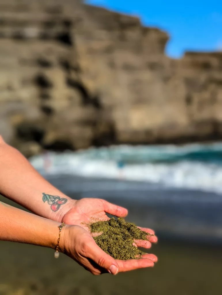 Une personne portant un bracelet et un tatouage tient du sable mouillé vert près du rivage sur Big Island