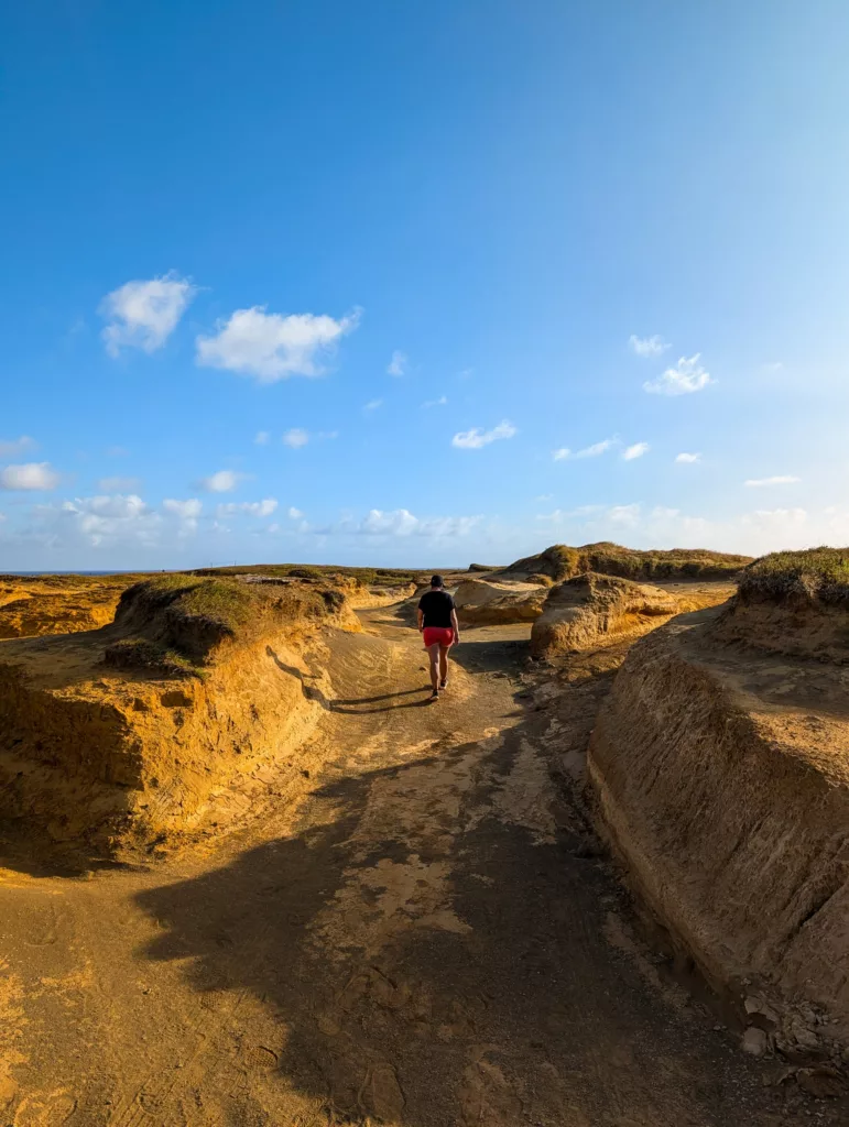 Une personne marche sur un chemin de terre entouré de collines sablonneuses et érodées, sous un ciel bleu vif parsemé de nuages