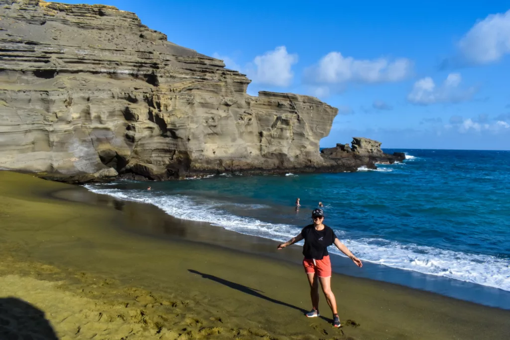 Une personne se tient sur une plage de sable verdâtre un site unique à Big Island, avec des falaises abruptes et des vagues bleues