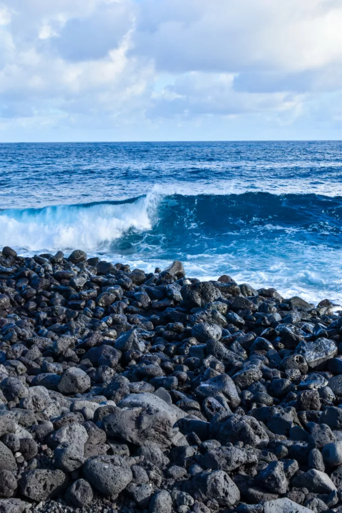 Un rivage avec des pierres volcaniques sombres au premier plan et une vague bleue de l'océan se brisant derrière elles