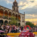 Une foule regarde un défilé de coccinelles Volkswagen vintage décorées dans une rue de Guadalajara, avec un bâtiment historique en pierre et un clocher au coucher du soleil