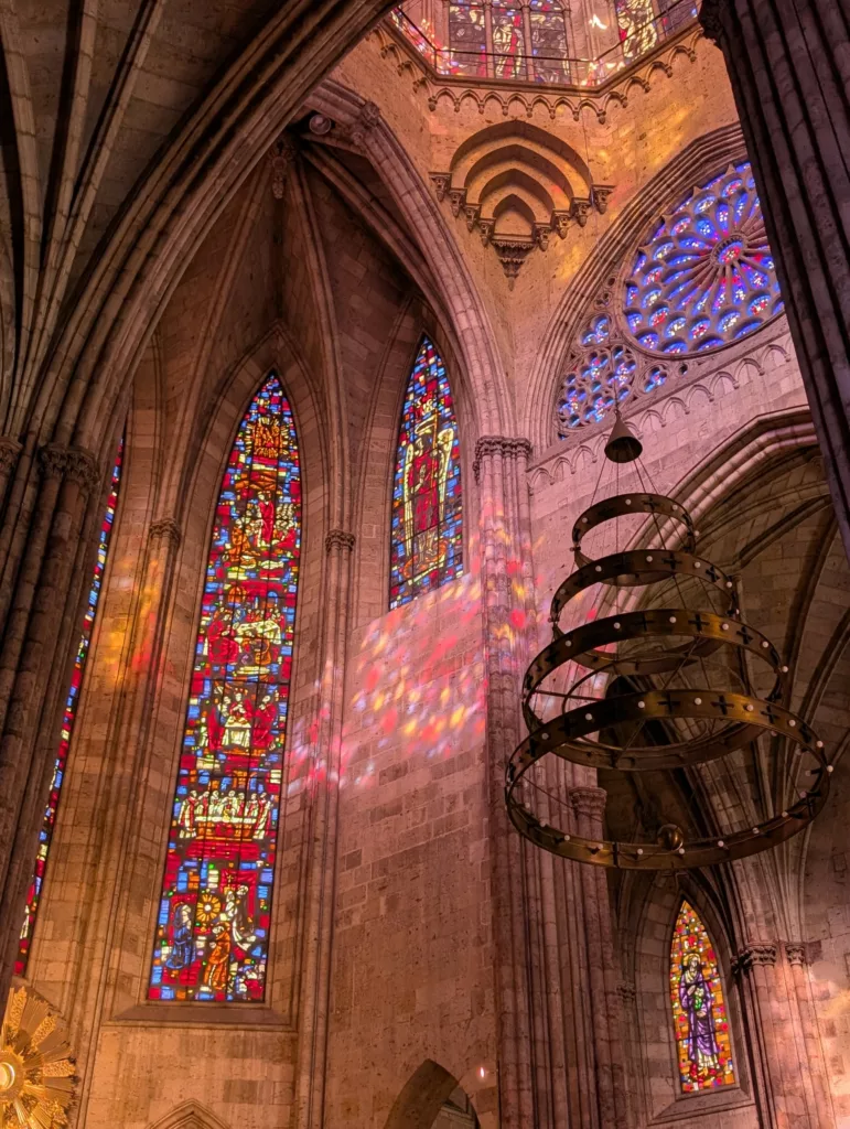De hauts vitraux et des rosaces illuminent l'intérieur en pierre de cette cathédrale gothique. La lumière projette des motifs sur les murs tandis qu'un grand lustre est suspendu au-dessus de la tête. 