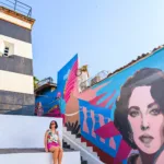 Une femme portant des lunettes de soleil et une tenue colorée est assise sur des marches blanches près d'une fresque murale