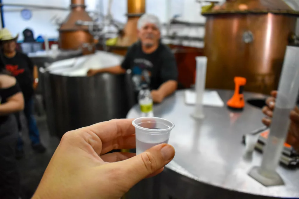 Une main tient un petit gobelet en plastique contenant un liquide transparent au premier plan,  et un grand équipement métallique à l'arrière-plan.