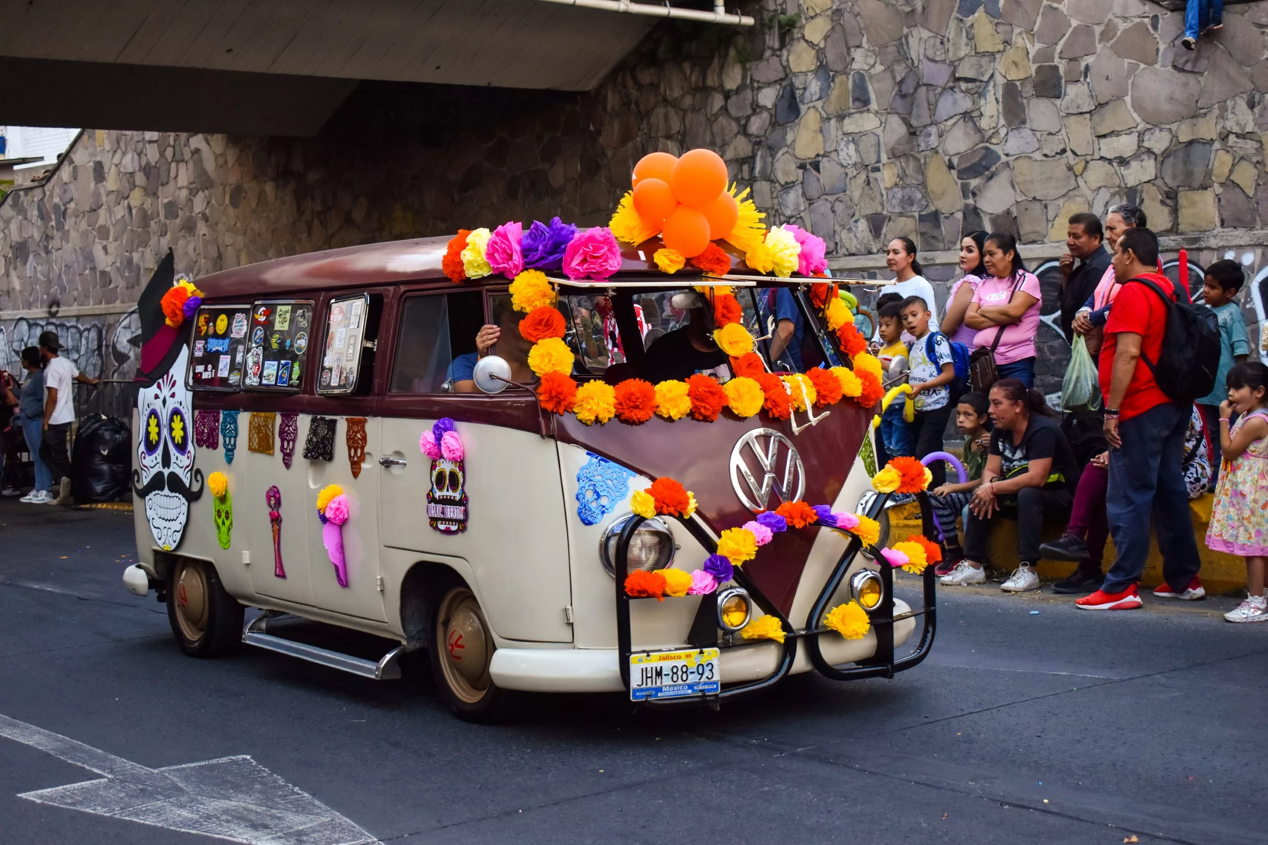 Un van Volkswagen vintage décorée de fleurs en papier et de ballons colorés descend une rue bordée de spectateurs
