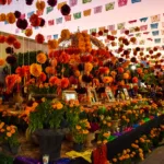 Un autel en plein air décoré de cempasúchil orange, de bougies, de photos encadrées et de banderoles colorées en papel picado suspendues au-dessus célèbre la fête des morts mexicaine, avec des personnes visibles à l'arrière-plan.