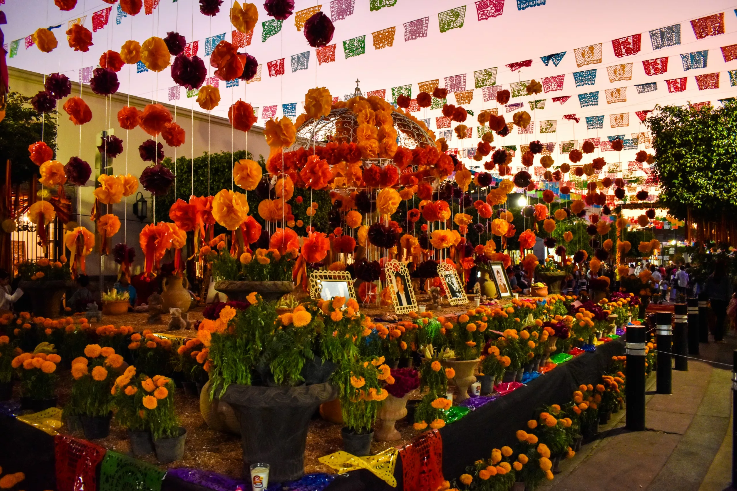 Un autel en plein air décoré de cempasúchil orange, de bougies, de photos encadrées et de banderoles colorées en papel picado suspendues au-dessus célèbre la fête des morts mexicaine, avec des personnes visibles à l'arrière-plan.