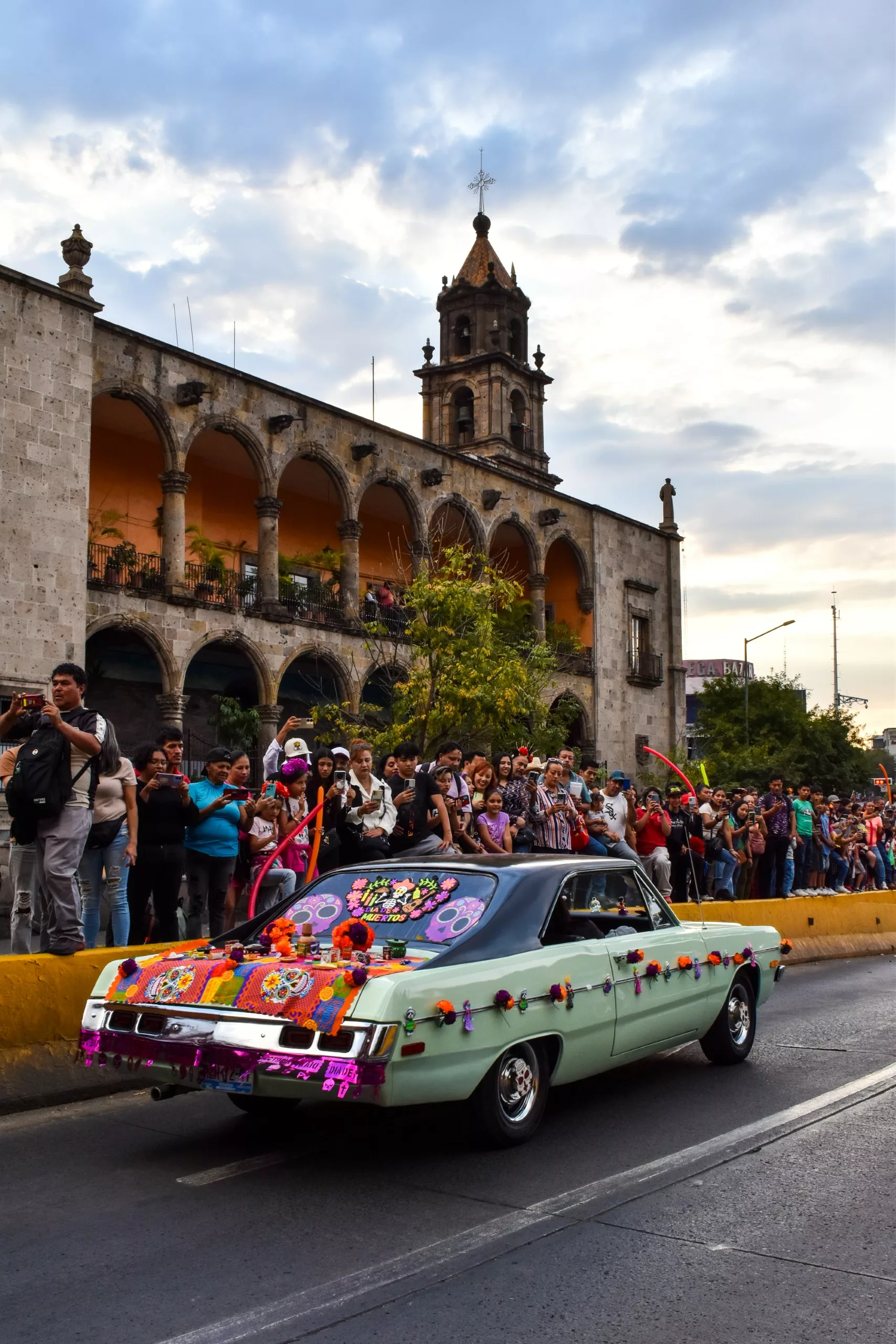 Une voiture ancienne décorée de fleurs colorées et de crânes pour la fête des morts mexicaine une rue de Guadalajara ; un bâtiment historique en pierre avec des arches et un clocher se trouve à l'arrière-plan.