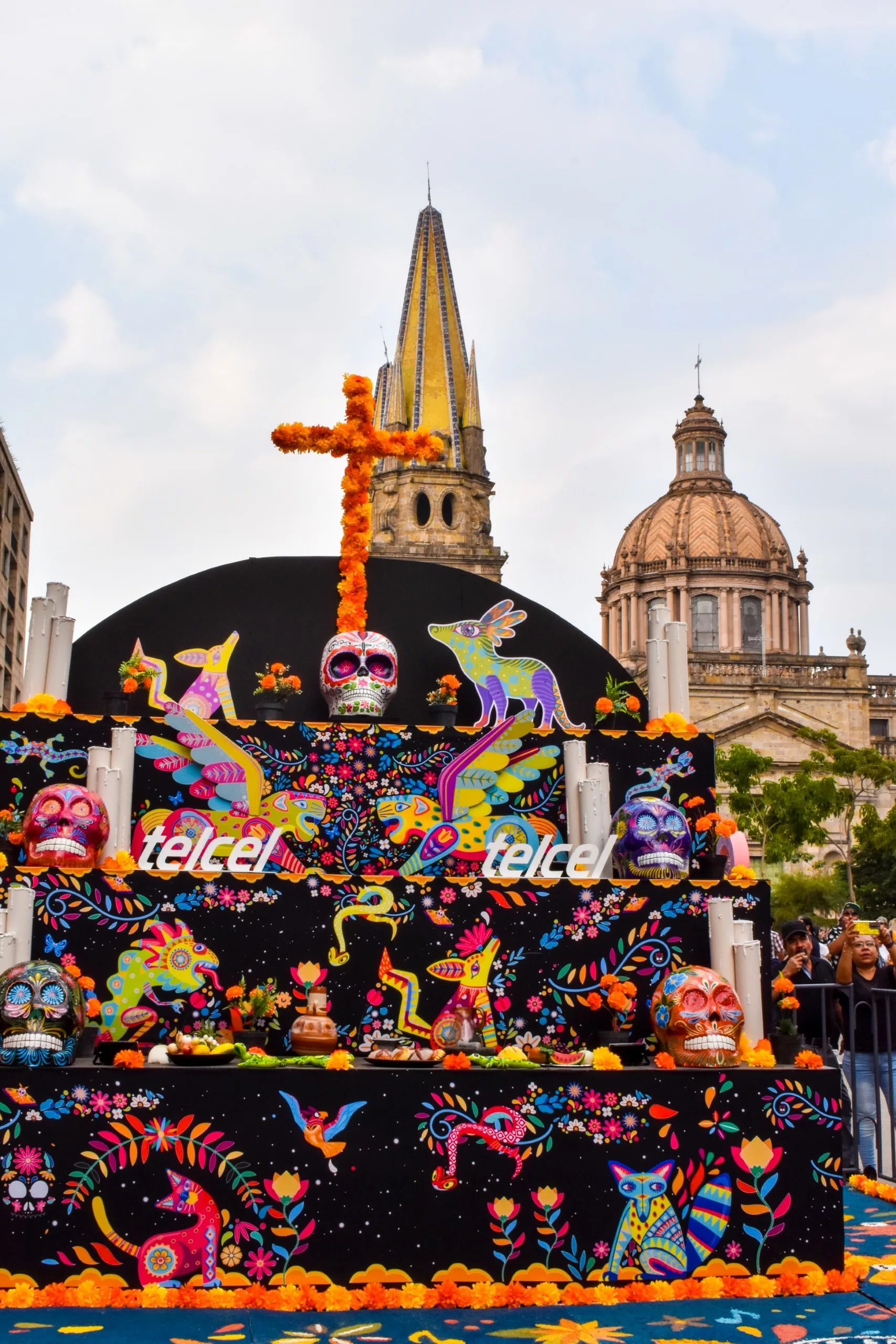 Un autel coloré du Jour des morts est orné de crânes peints, de figures animales vibrantes et de fleurs devant des bâtiments historiques et une église à la haute flèche.