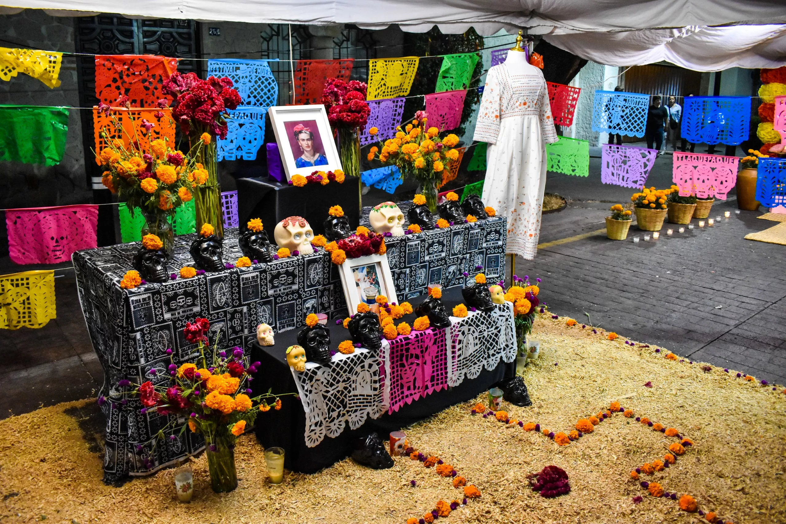 Un vibrant autel de la fête des morts orné de fleurs, de papel picado, de bougies, de crânes, d'une photo de Frida Kahlo, de vases, d'une robe blanche et de compositions florales colorées sur le sol.