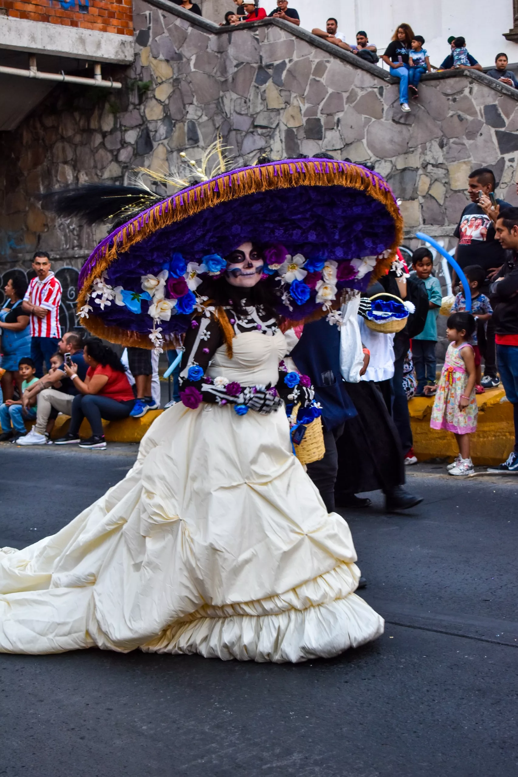 Une femme vêtue d'une grande robe blanche et d'un chapeau surdimensionné décoré de fleurs violettes et bleues participe au défilé de la fête des morts mexicaine, le visage peint en forme de crâne.