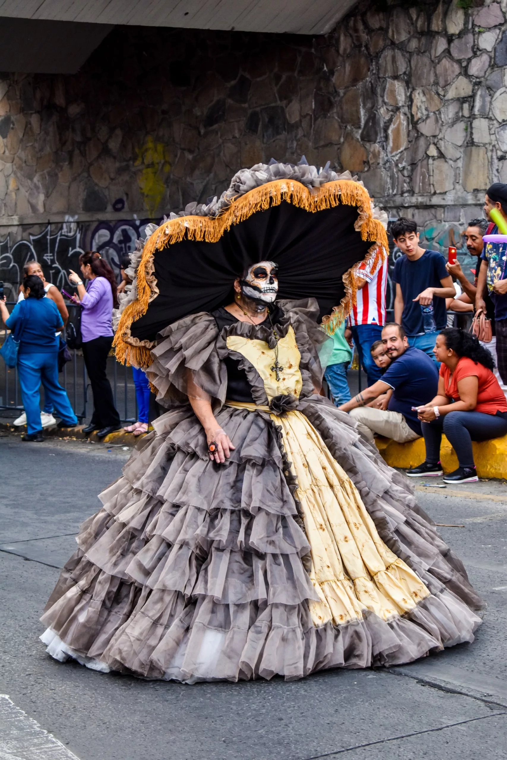 Une personne portant une grande robe grise et beige à volants, un chapeau à larges bords avec des bordures dorées et une peinture pour visage de squelette marche dans un défilé célébrant la fête des morts au Mexique