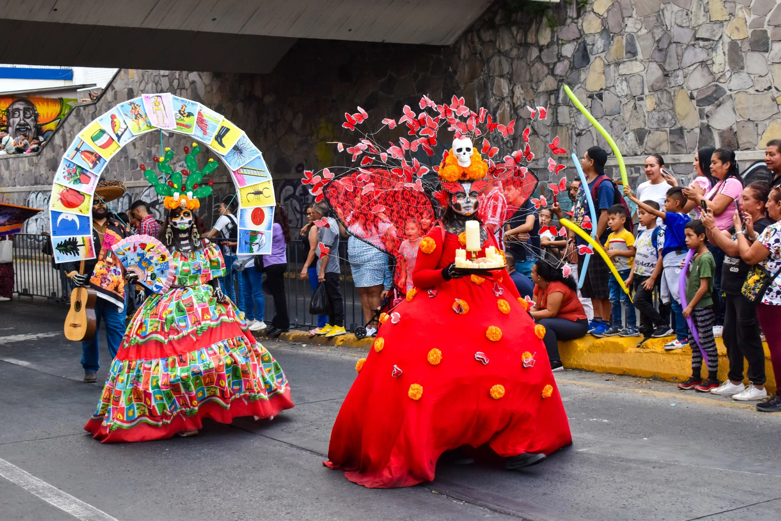 Deux personnes habillées en Catrinas colorées marchent dans un défilé de la fête des morts mexicaine ; l'une porte une robe multicolore, l'autre est vêtue de rouge avec une bougie et un couvre-chef en forme de tête de mort.