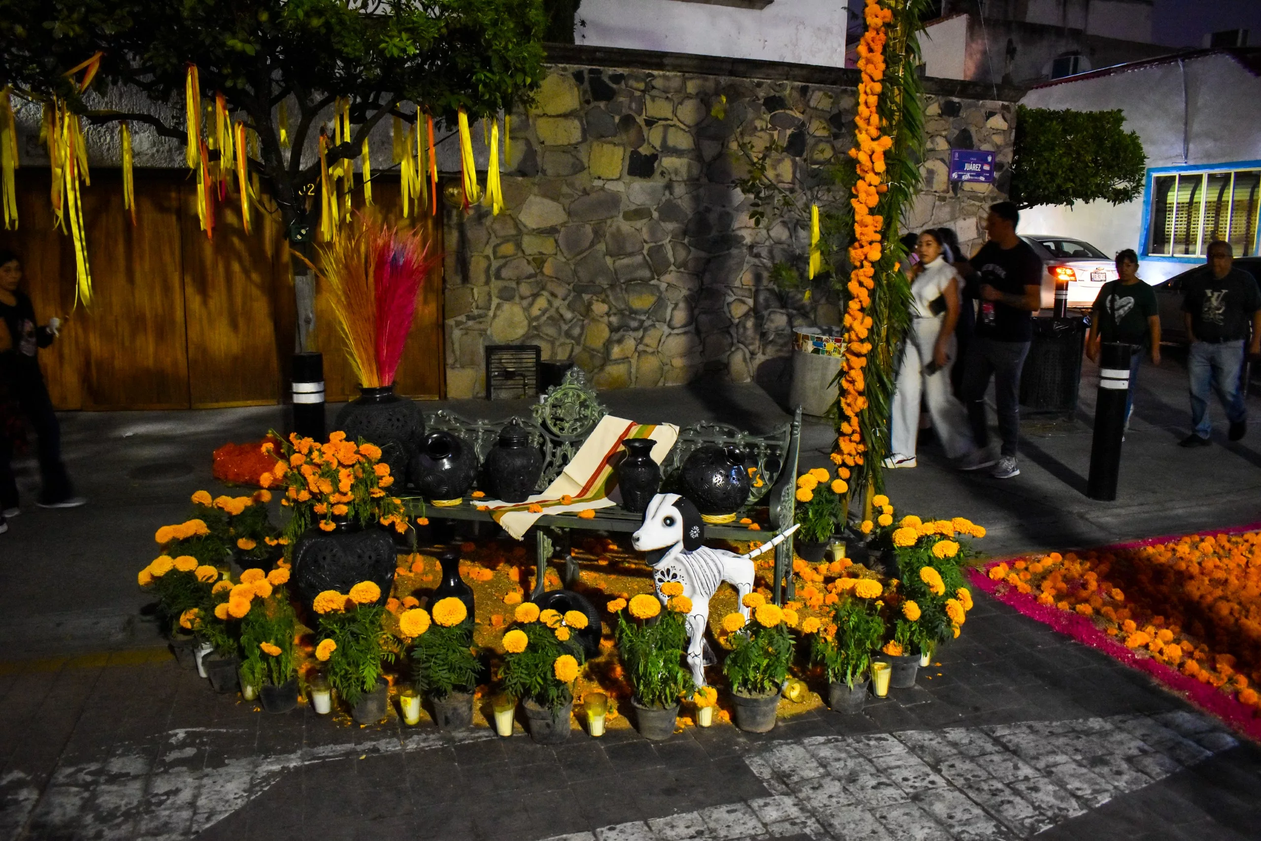 Un autel du Día de Muertos orné de fleurs, de crânes, de bougies, de rubans jaunes dans un arbre et d'un petit chien squelettique
