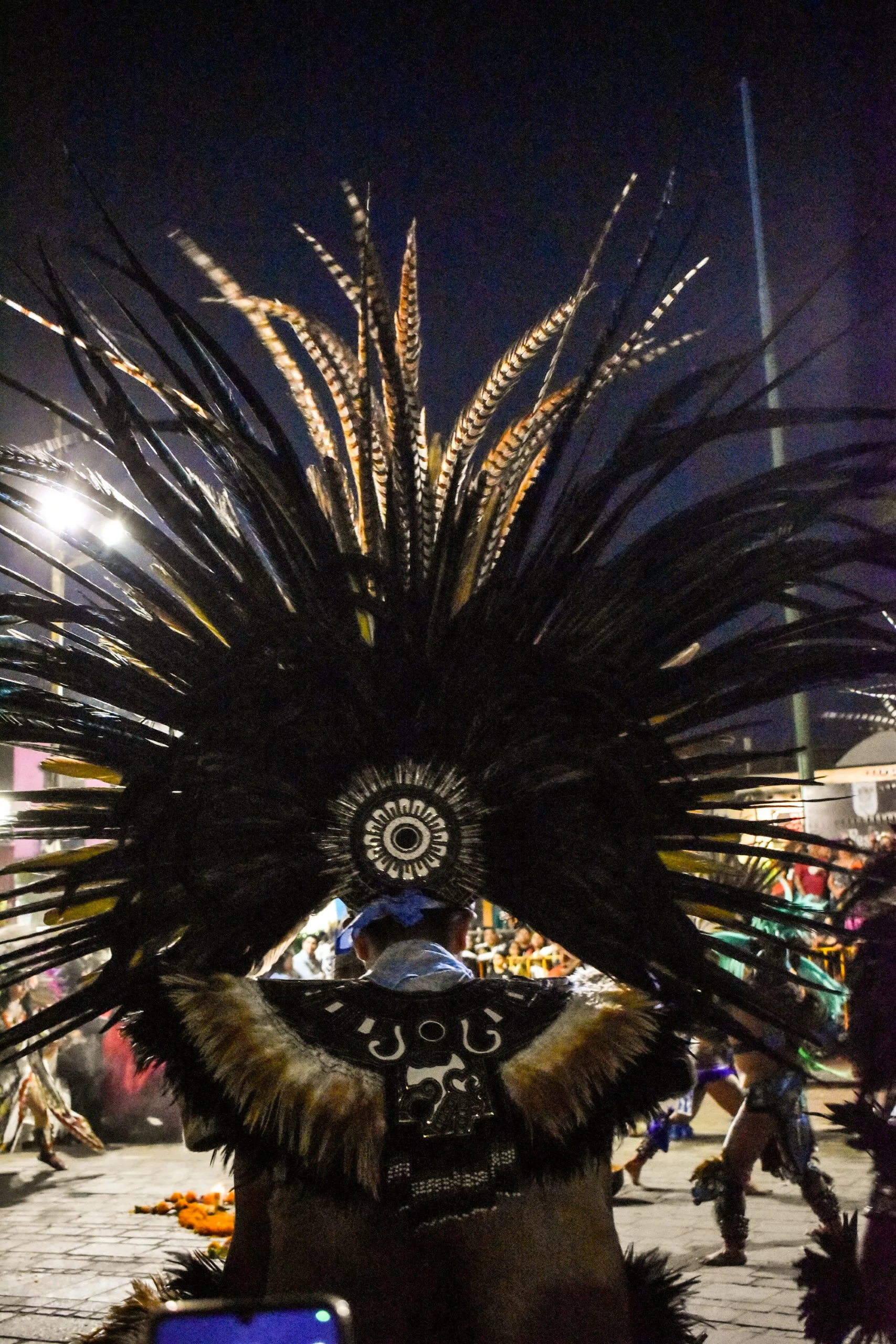 Une personne portant une grande coiffe à plumes et des vêtements traditionnels se tient à l'extérieur, la nuit