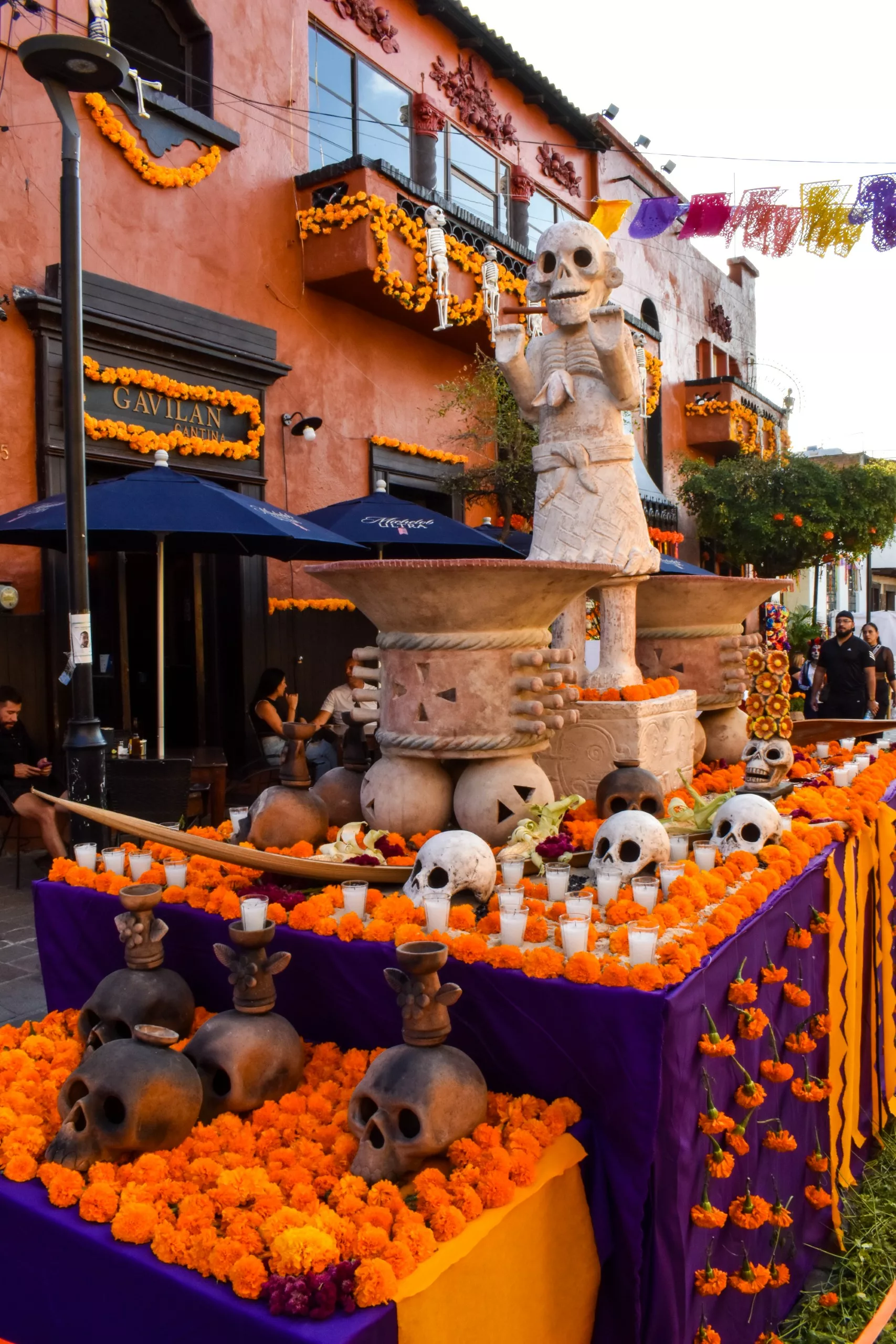 Un grand autel pour la fête des morts mexicaine, orné de fleurs de cempasúchil, de bougies et de sculptures de crânes, se dresse dans une rue. Une grande statue de squelette est la pièce maîtresse au milieu de bâtiments orange et de banderoles en papel picado.