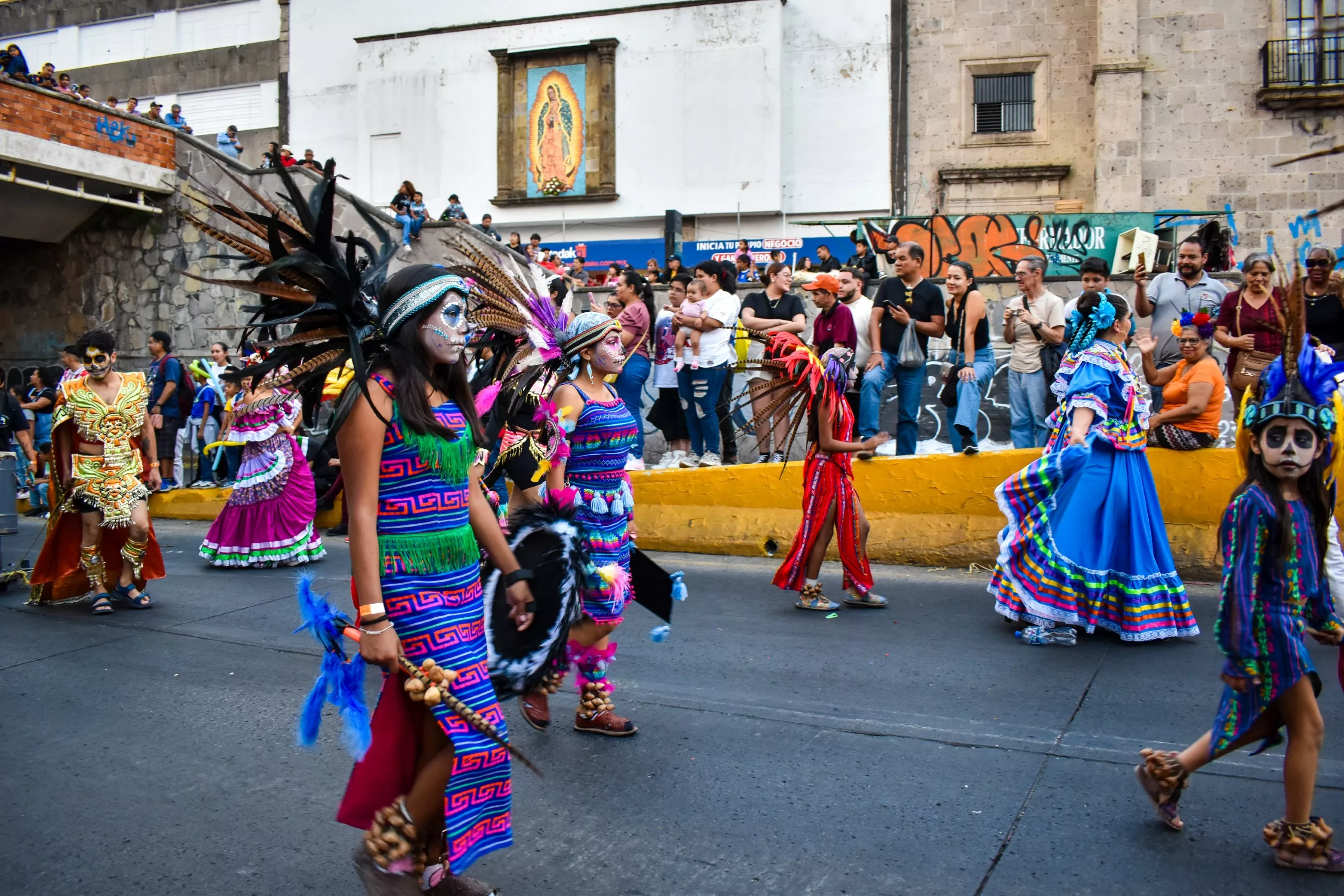 Des personnes vêtues de costumes aztèques traditionnels colorés et de coiffes à plumes défilent , sous le regard des spectateurs derrière une barrière jaune. Une peinture murale de la Vierge de Guadalupe orne un bâtiment voisin.