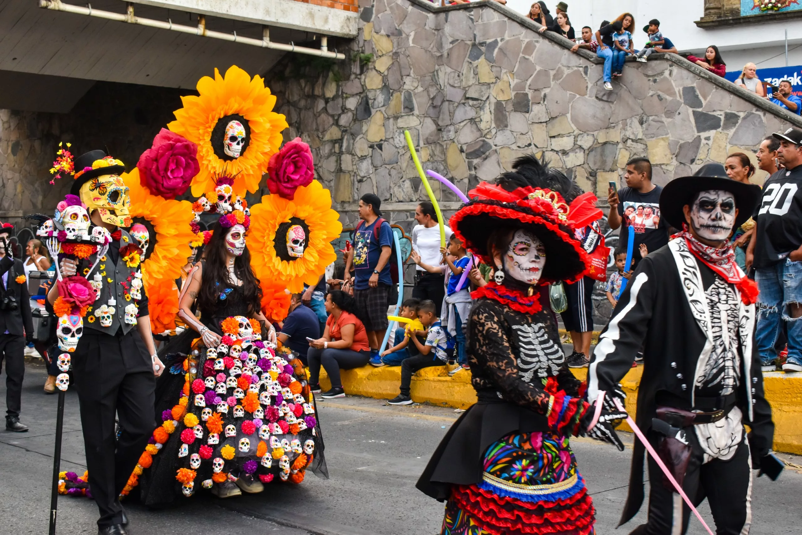Un groupe de personnes en costumes de la fête des morts mexicaine et maquillées marchent dans un défilé. L'une d'entre elles porte une robe noire avec des fleurs et de grandes ailes de fleurs orange