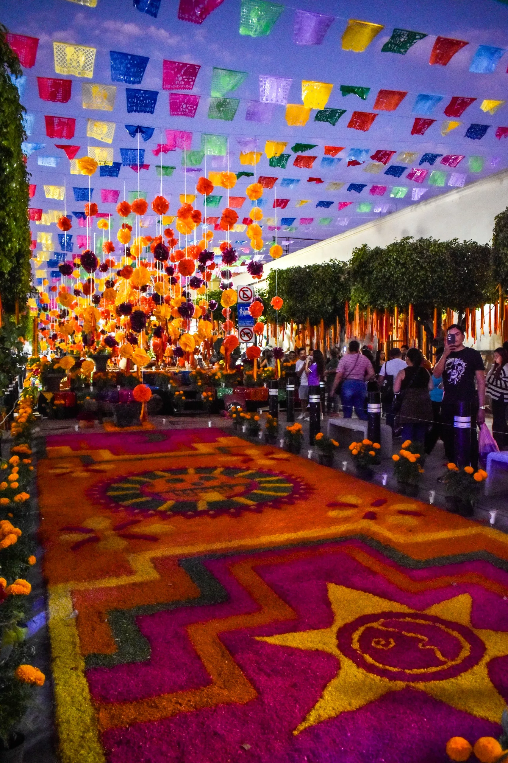 Une allée décorée de banderoles en papier colorées, de cempasúchil et de tapis de fleurs vibrantes célèbre l'esprit festif de la fête des morts mexicaine. Les gens se promènent sur le chemin bordé d'arbres