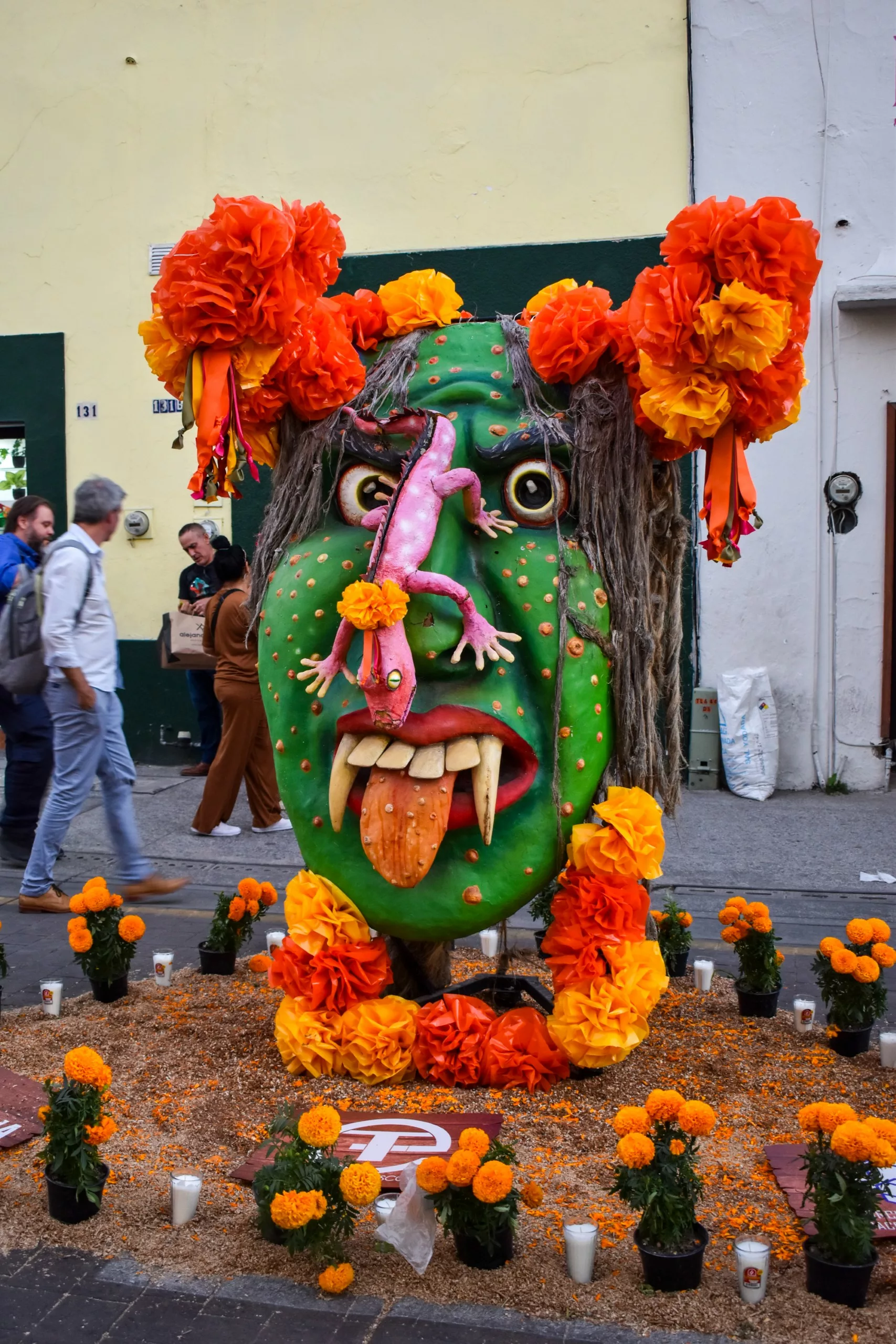 Une grande sculpture colorée pour la fête des morts mexicaine représente un visage vert avec des crocs, une langue large et un lézard rose sur le nez, orné de fleurs et entouré de bougies