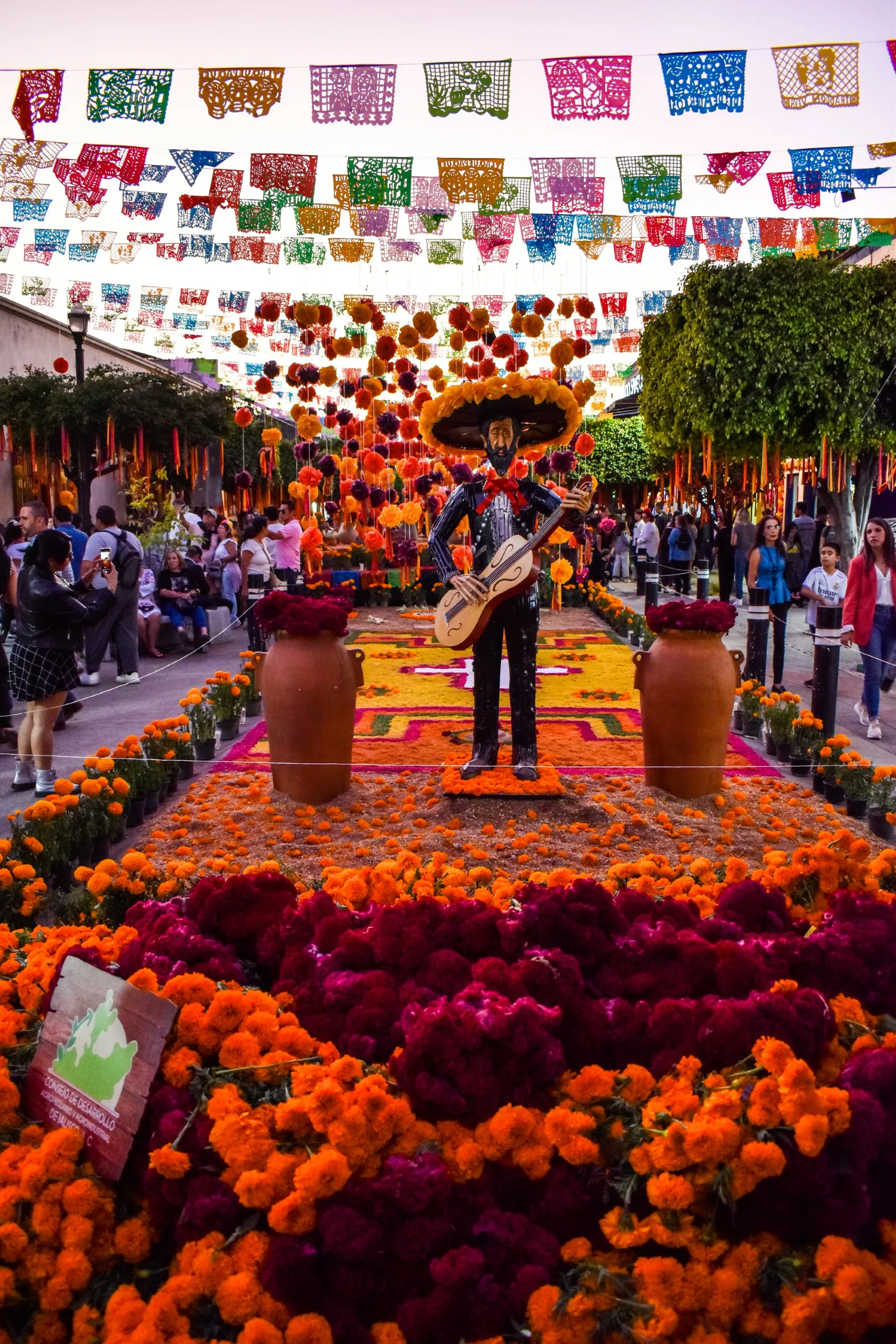 Une rue avec de nombreuses fleurs, de papel picado coloré et d'une statue de mariachi avec une guitare