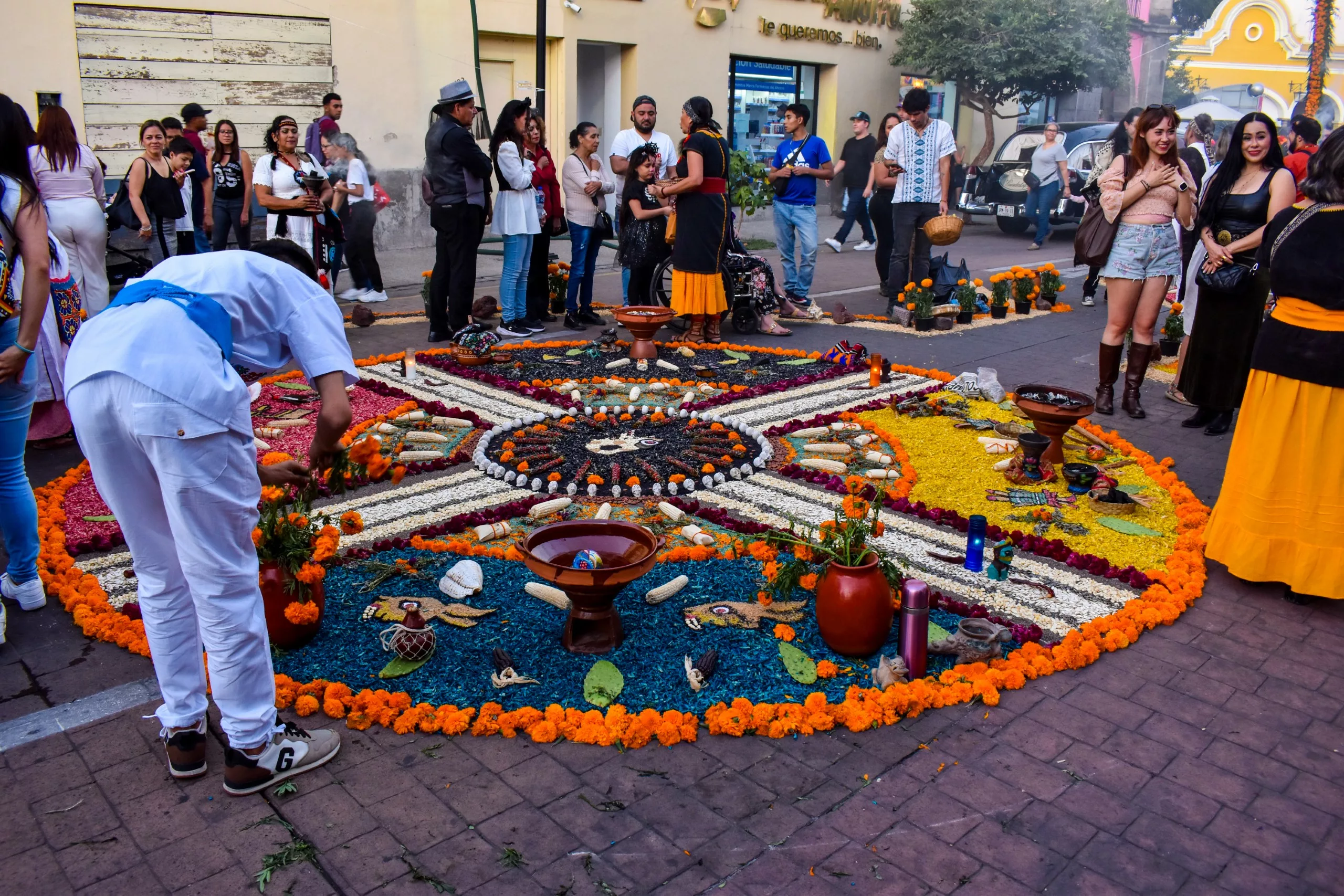 Les gens se rassemblent autour d'un grand tapis de fleurs coloré du Jour des morts, avec des motifs complexes de pétales, de bougies et d'offrandes. Certains participants portent des costumes et des vêtements traditionnels.