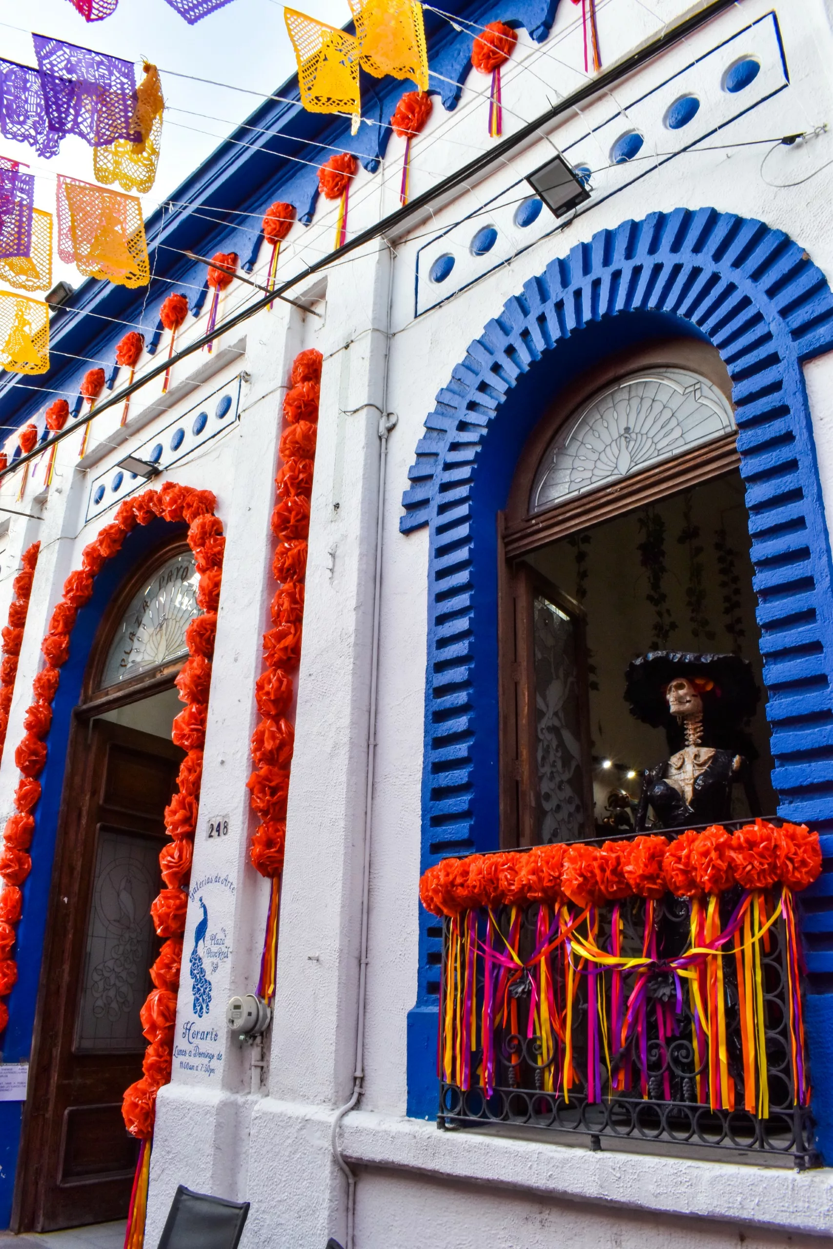 Un bâtiment décoré pour la fête des morts mexicains, avec des cempasúchil orange, des bannières en papier colorées, des rubans rouges et jaunes, et une statue d'un squelette en costume mexicain traditionnel visible à travers une fenêtre aux arches bleues.