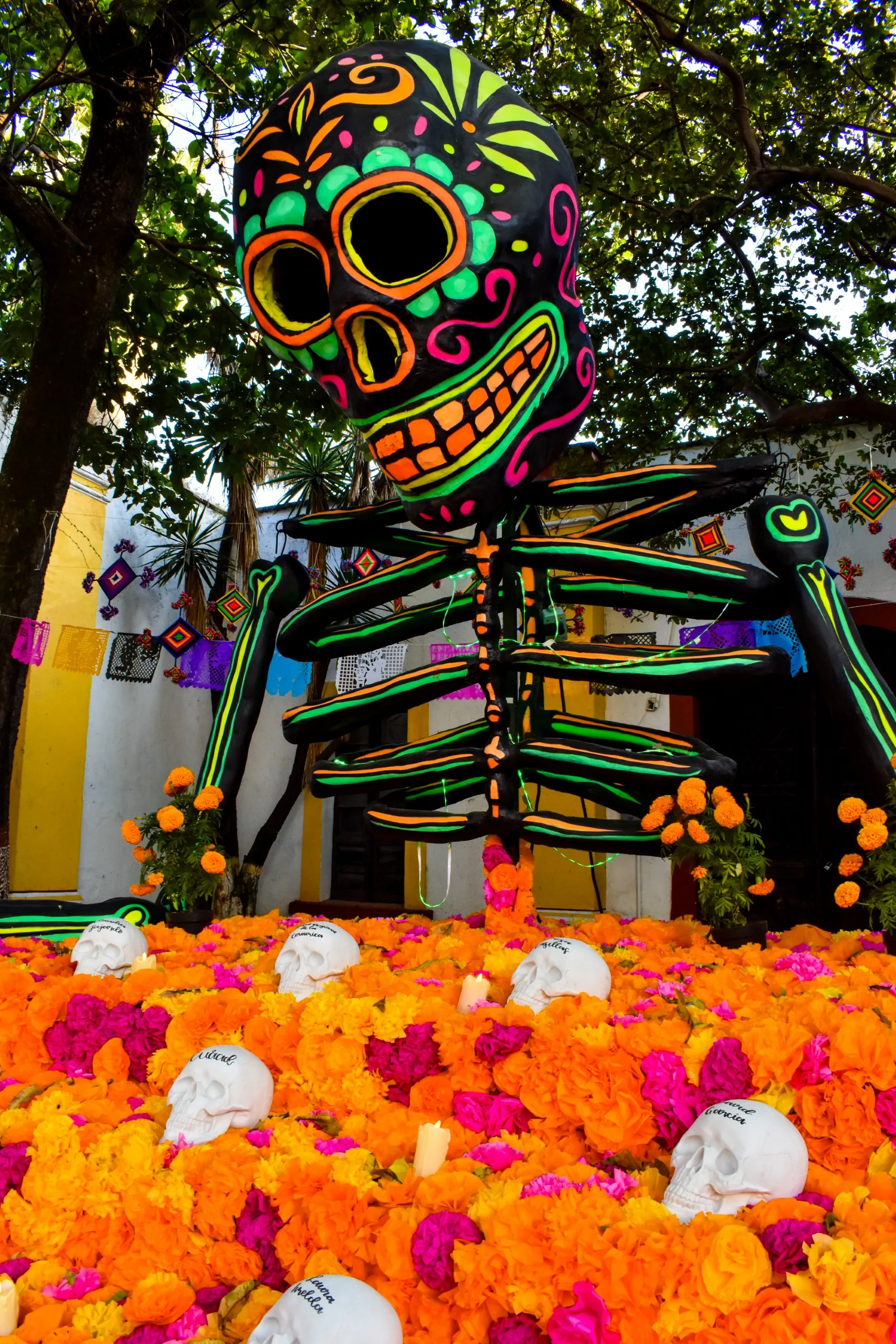 Un grand squelette coloré, décoré de motifs au néon, se tient derrière un autel de soucis lumineux et de crânes blancs, célébrant le Dia de Muertos mexicain en plein air, sous des arbres.