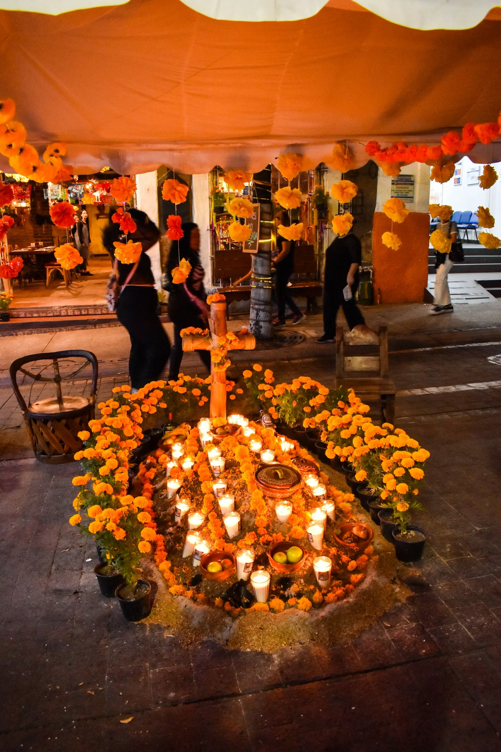 Un autel du Día de Muertos est orné de fleurs, de bougies et d'offrandes sous un dais, tandis que des gens passent à l'arrière-plan.