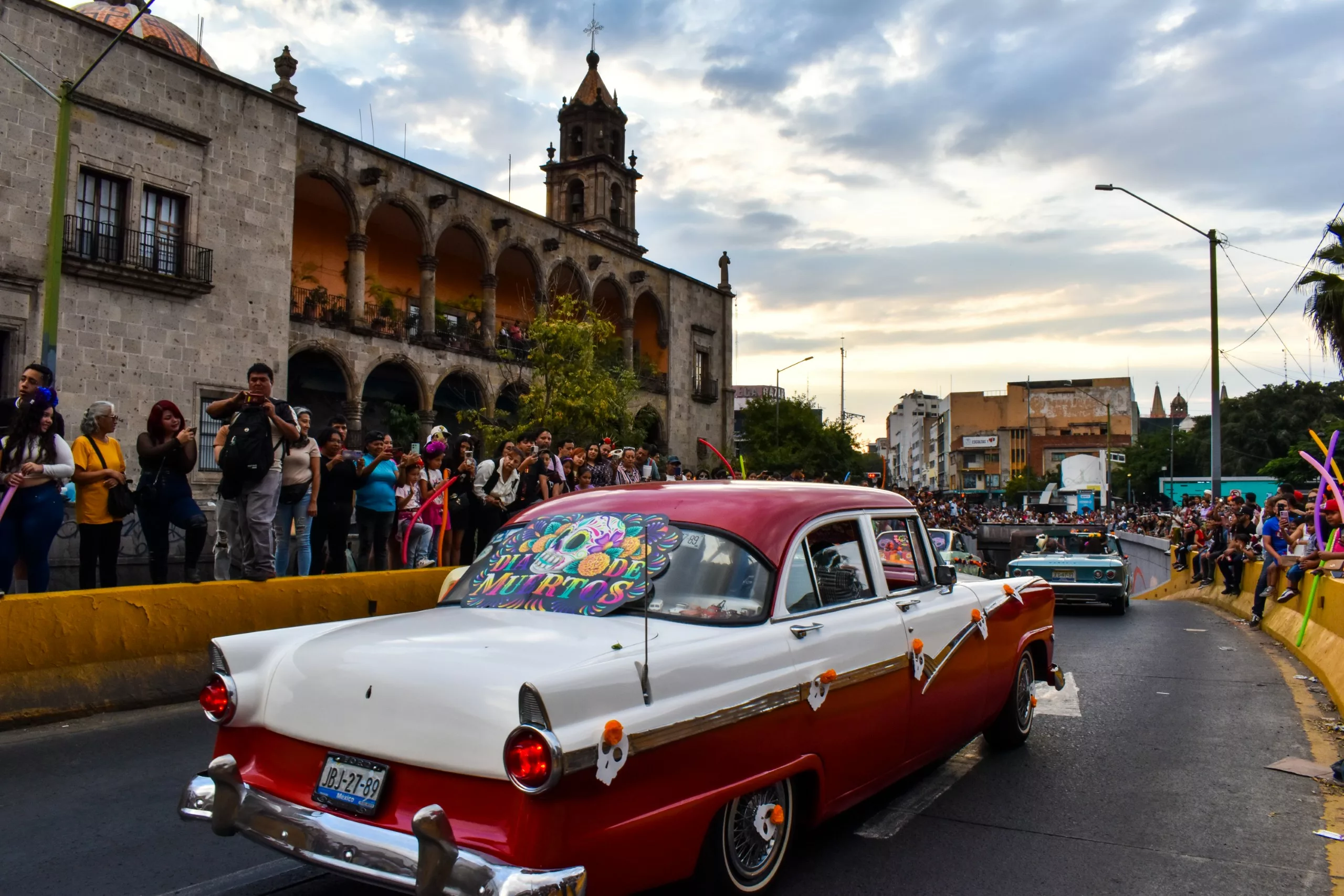 Une voiture ancienne rouge et blanche décorée de fleurs descend une rue lors d'un défilé de la fête des morts mexicaine, sous le regard de la foule depuis le trottoir. Un vieux bâtiment en pierre et le clocher d'une église se dressent à l'arrière-plan.