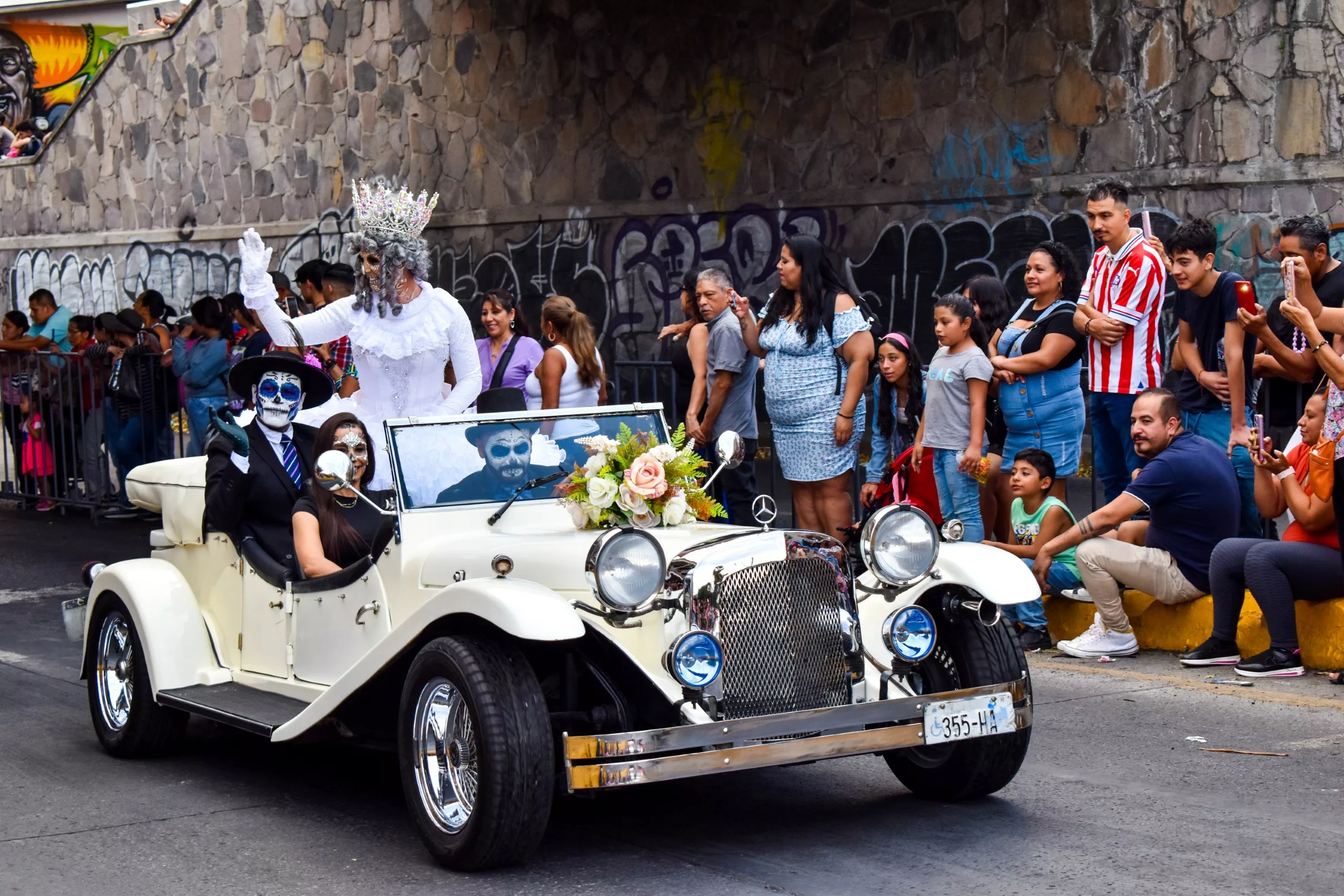 Une voiture de collection décorée, avec des personnes en costumes de squelette, roule dans un défilé de la fête des morts au Mexique, tandis qu'une figure couronnée en blanc se tient à l'arrière