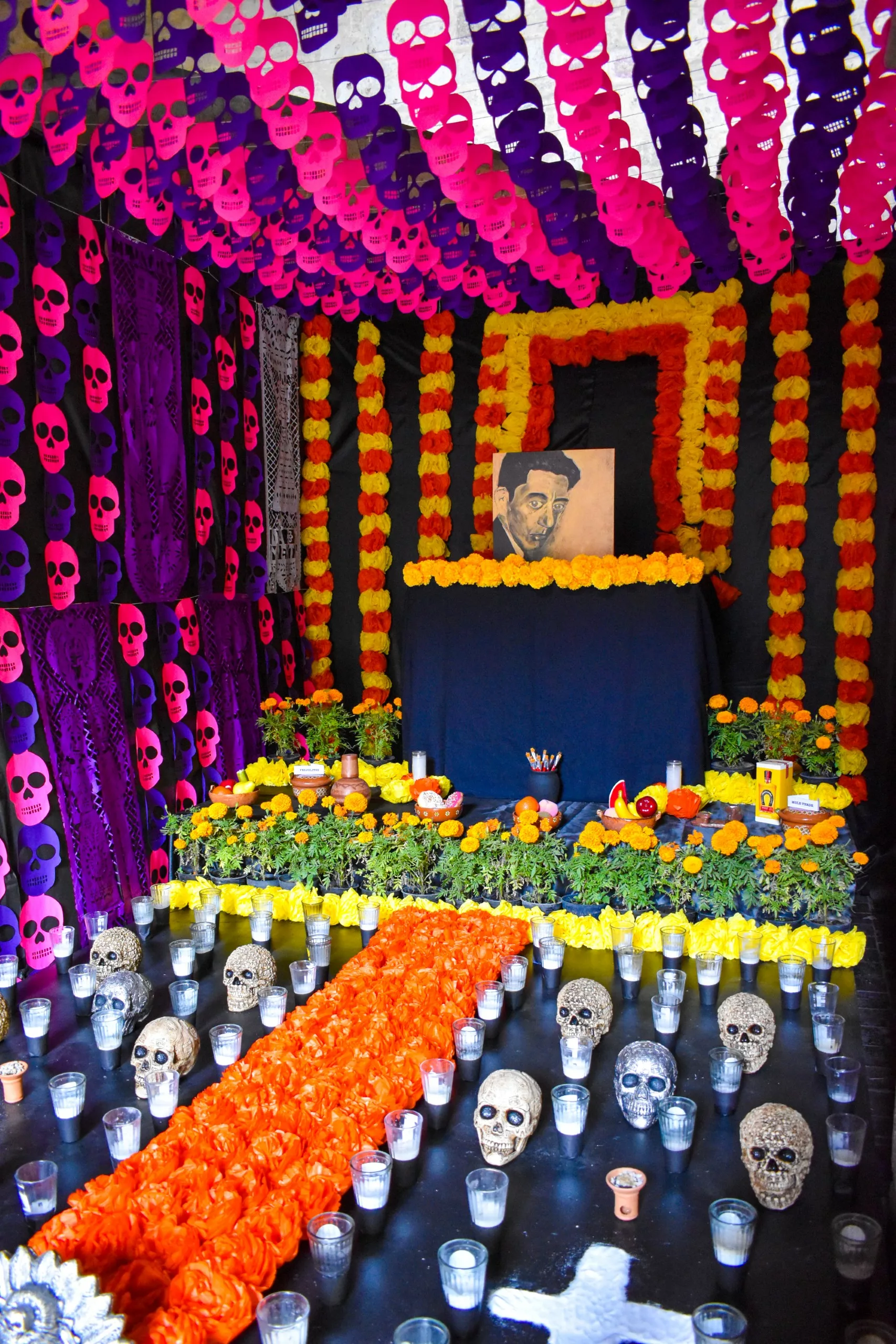 Autel décoré pour le Día de los Muertos, ou fête des morts mexicaine, avec des fleurs de cempasúchil, des bougies, des crânes, des bannières en papel picado, des offrandes et un portrait central sur fond noir et jaune.
