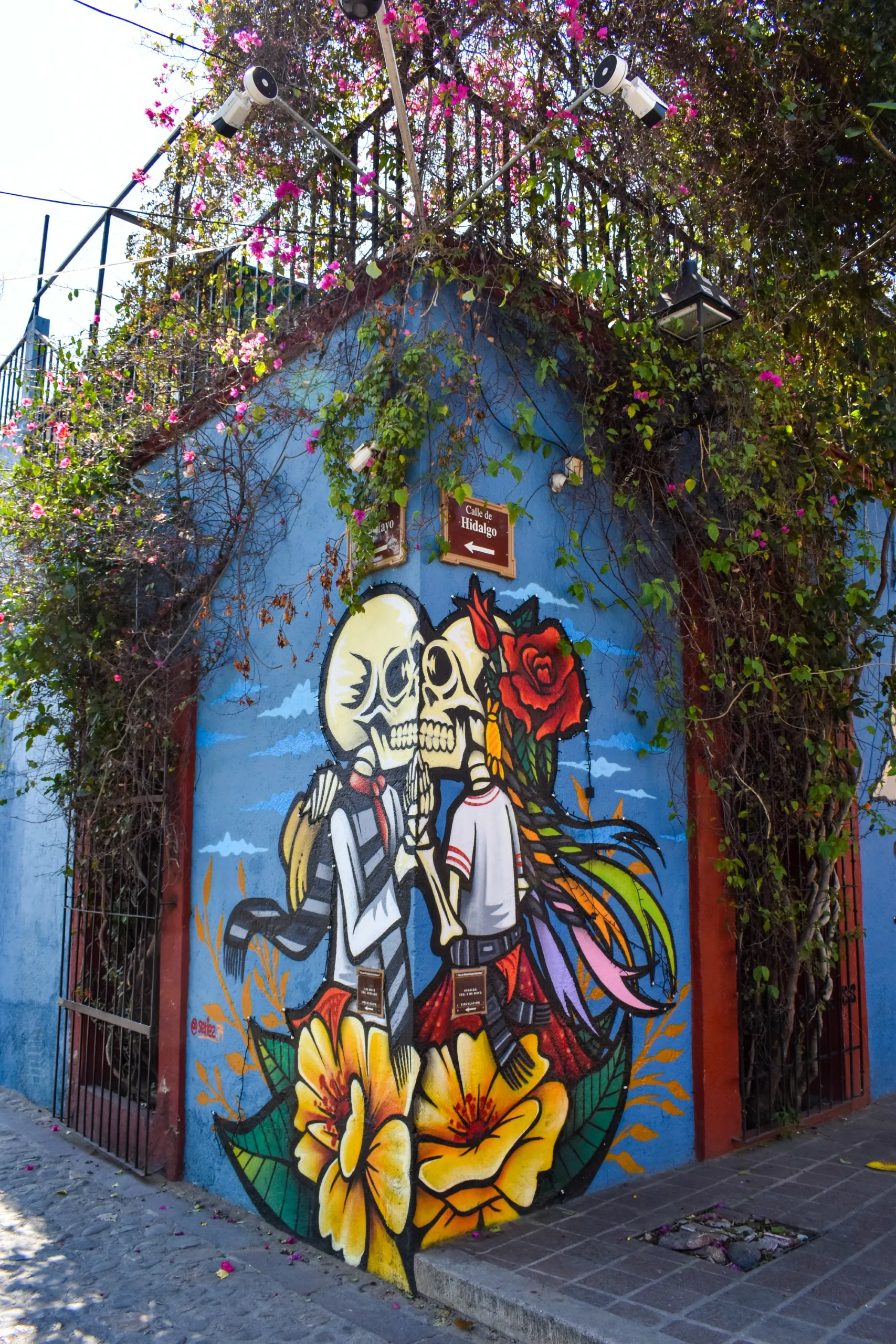 Une peinture murale colorée célébrant montre deux squelettes en vêtements traditionnels, entourés de fleurs et d'une rose rouge sur un bâtiment bleu recouvert de vigne et de fleurs roses.