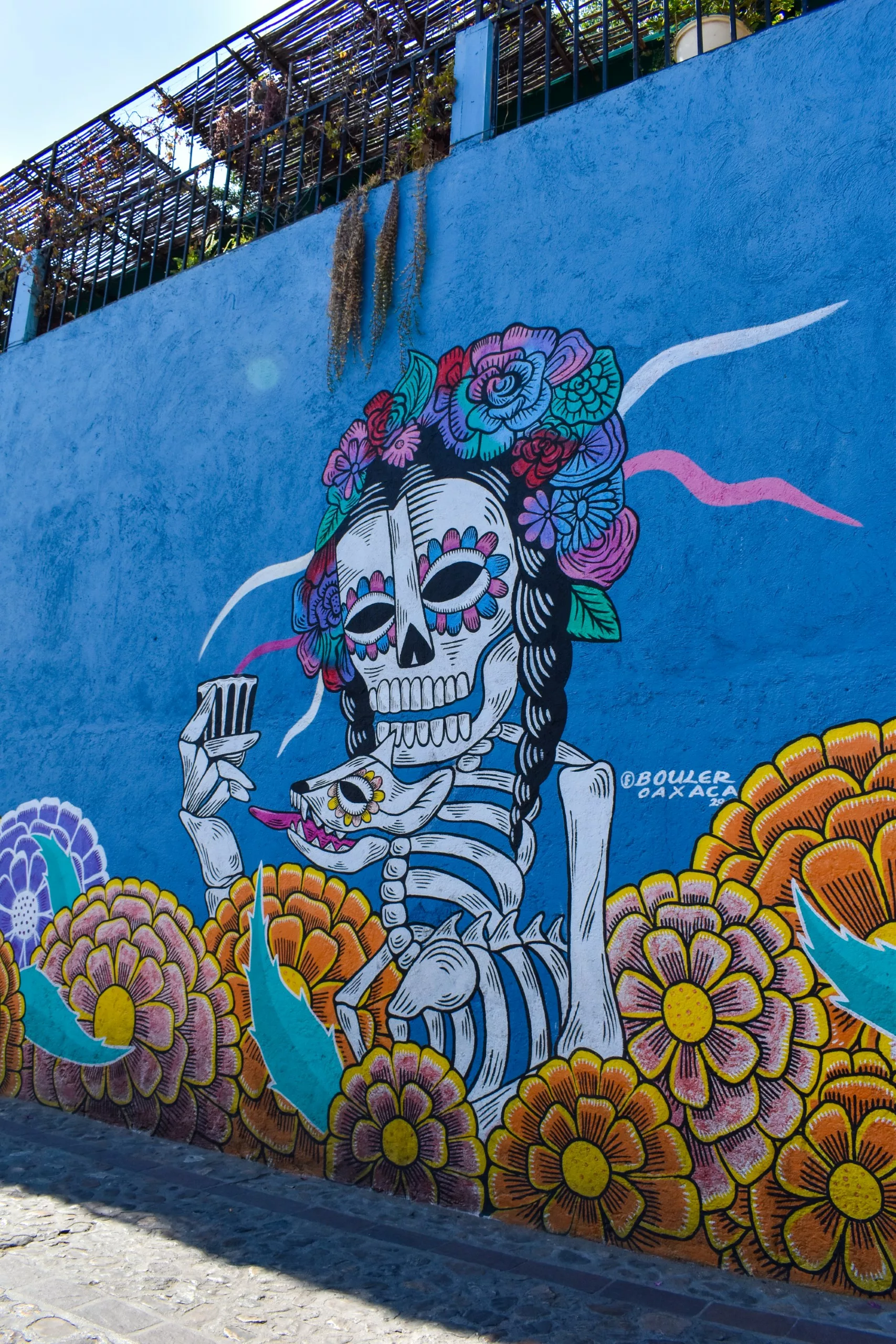 Une peinture murale colorée sur un mur bleu représente un squelette aux cheveux tressés et à la coiffe florale, entouré de fleurs et de feuilles éclatantes. La peinture murale est signée "Bauer Oaxaca".