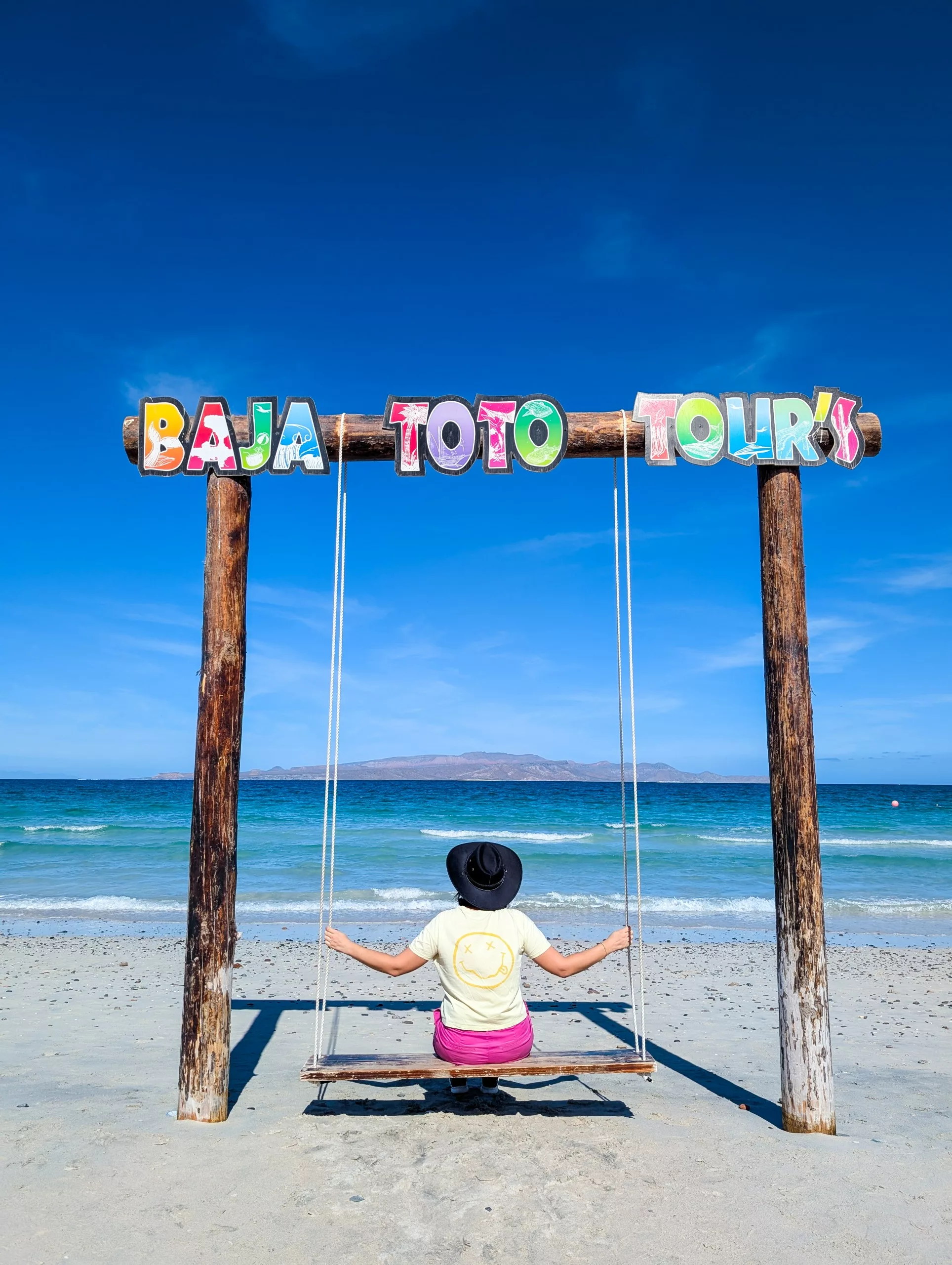 Une personne portant une chemise jaune et un chapeau noir est assise sur une balançoire sous un panneau "Baja Toto Tours", regardant l'océan.