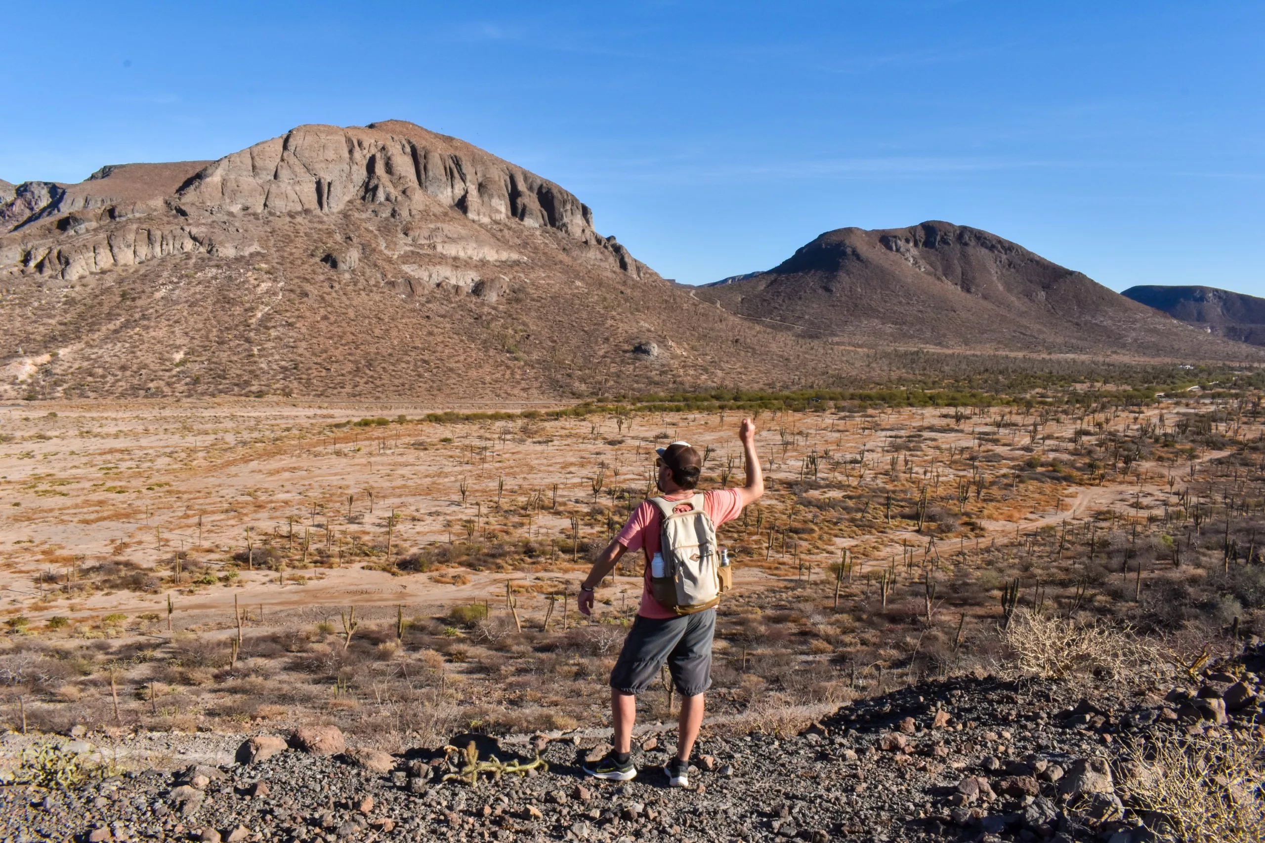 Une personne portant un sac à dos se tient sur un terrain rocailleux face à un paysage désertique sec avec une végétation clairsemée et deux grandes collines escarpées.