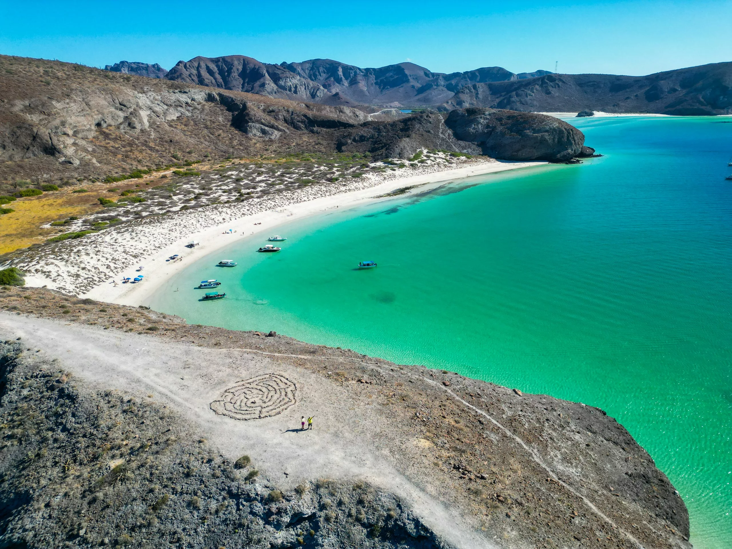Vue aérienne d'une baie incurvée avec une eau turquoise et une plage de sable blanc. De petits bateaux sont ancrés sur les rochers, et un dessin en spirale est tracé dans la terre au sommet de la falaise qui surplombe le rivage.