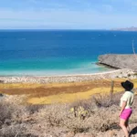 Une personne portant un chapeau et un short rose se tient sur une colline sèche et rocheuse surplombant une plage de sable et une eau turquoise