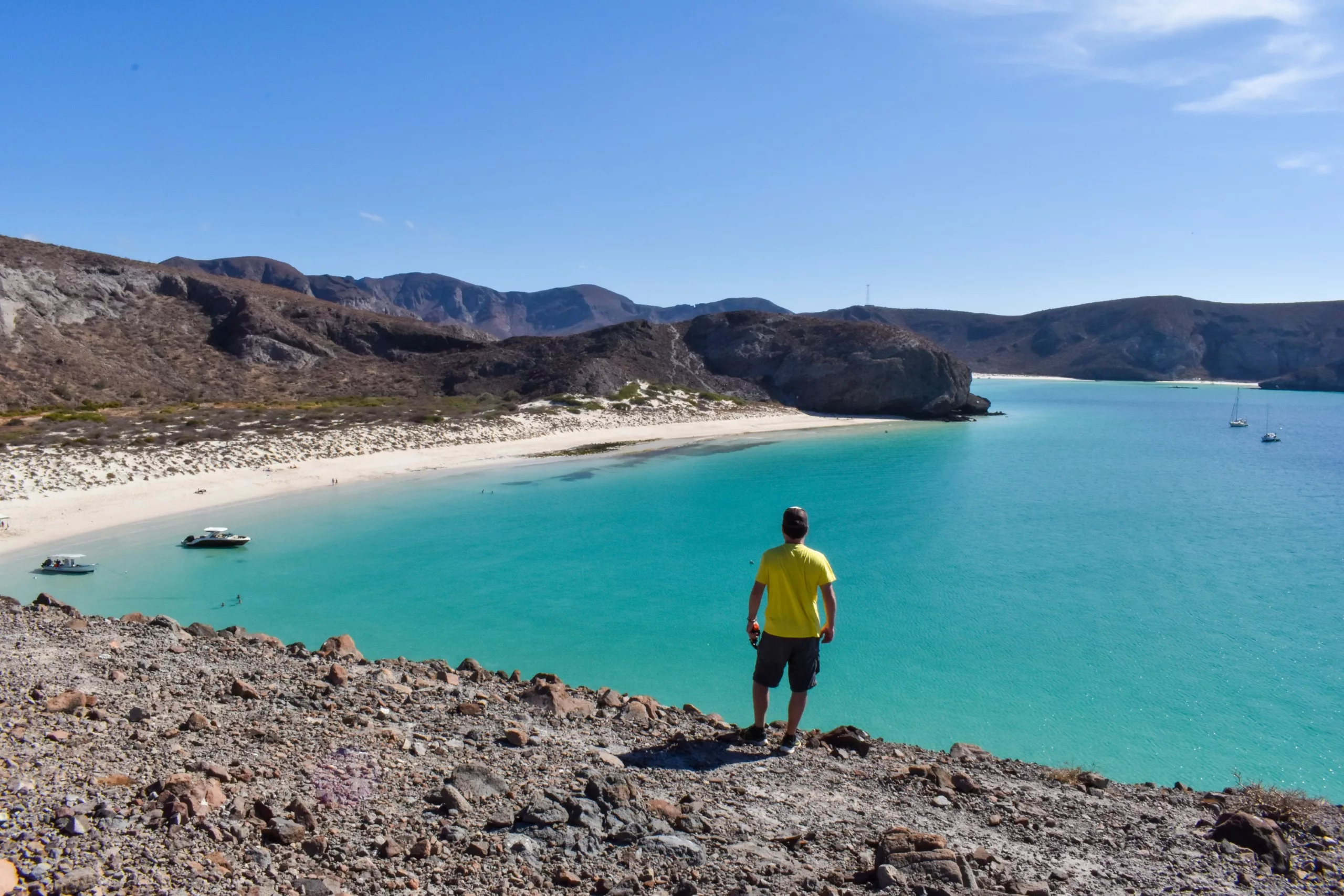 Une personne se tient sur une colline rocheuse surplombant la baie turquoise de La Paz, au Mexique, avec une plage de sable, un ciel bleu clair, des bateaux ancrés et des collines escarpées en arrière-plan.