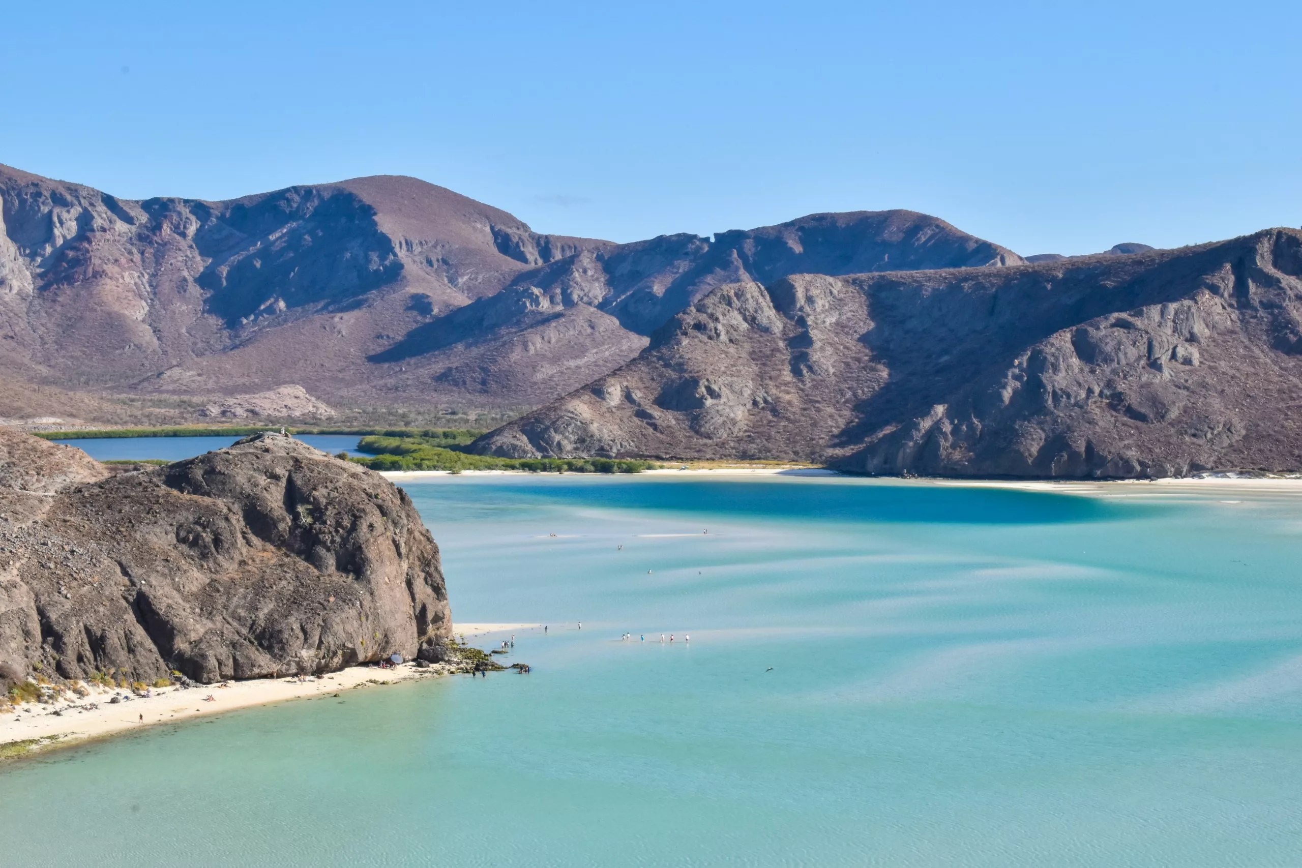 Une large baie turquoise et limpide est bordée de rivages sablonneux et de montagnes brunes et escarpées, sous un ciel bleu. Quelques petites silhouettes sont visibles sur l'eau et sur la plage.
