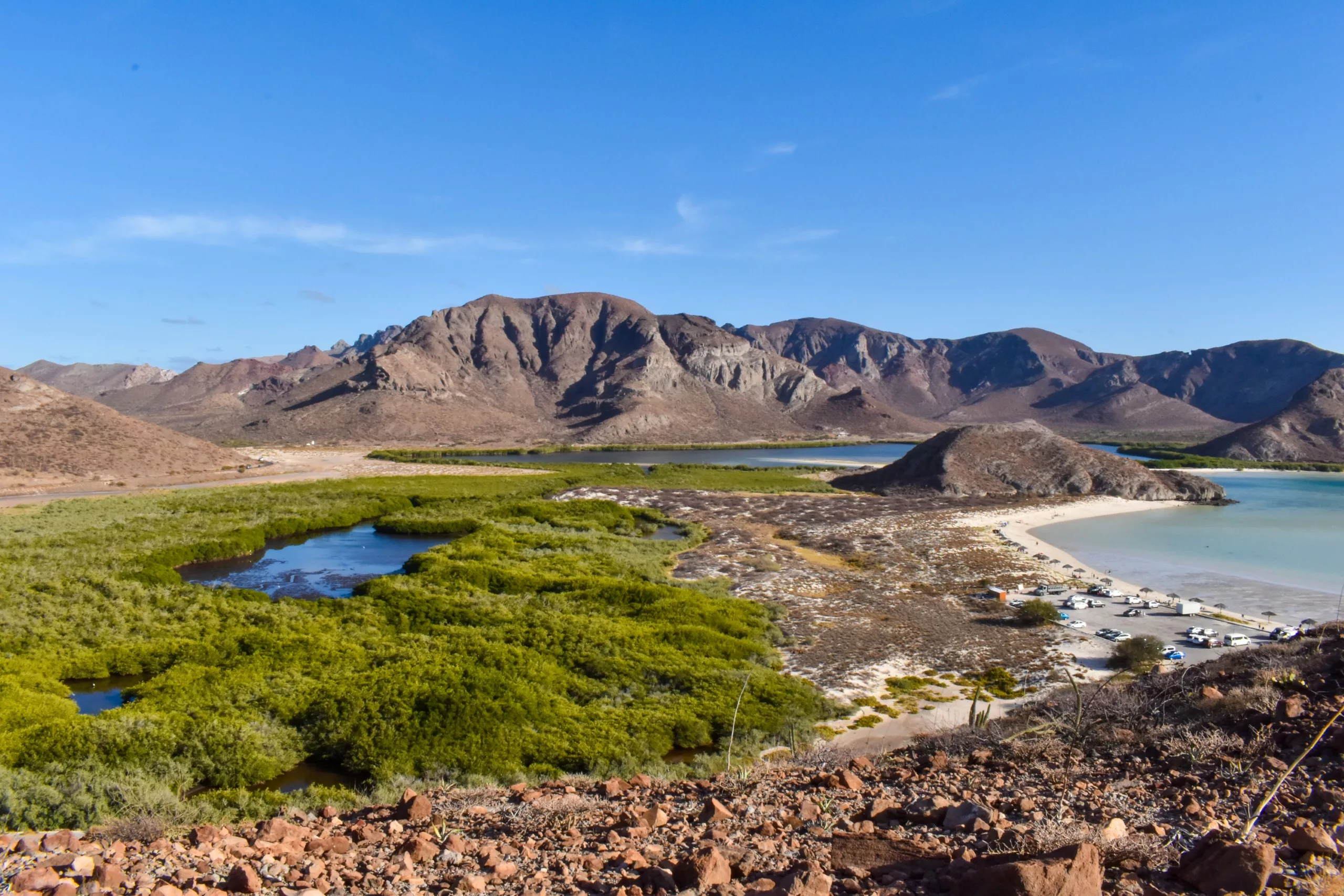 Paysage côtier avec des collines rocheuses en arrière-plan, de la végétation verte et de l'eau en avant-plan, une plage de sable avec des voitures garées.