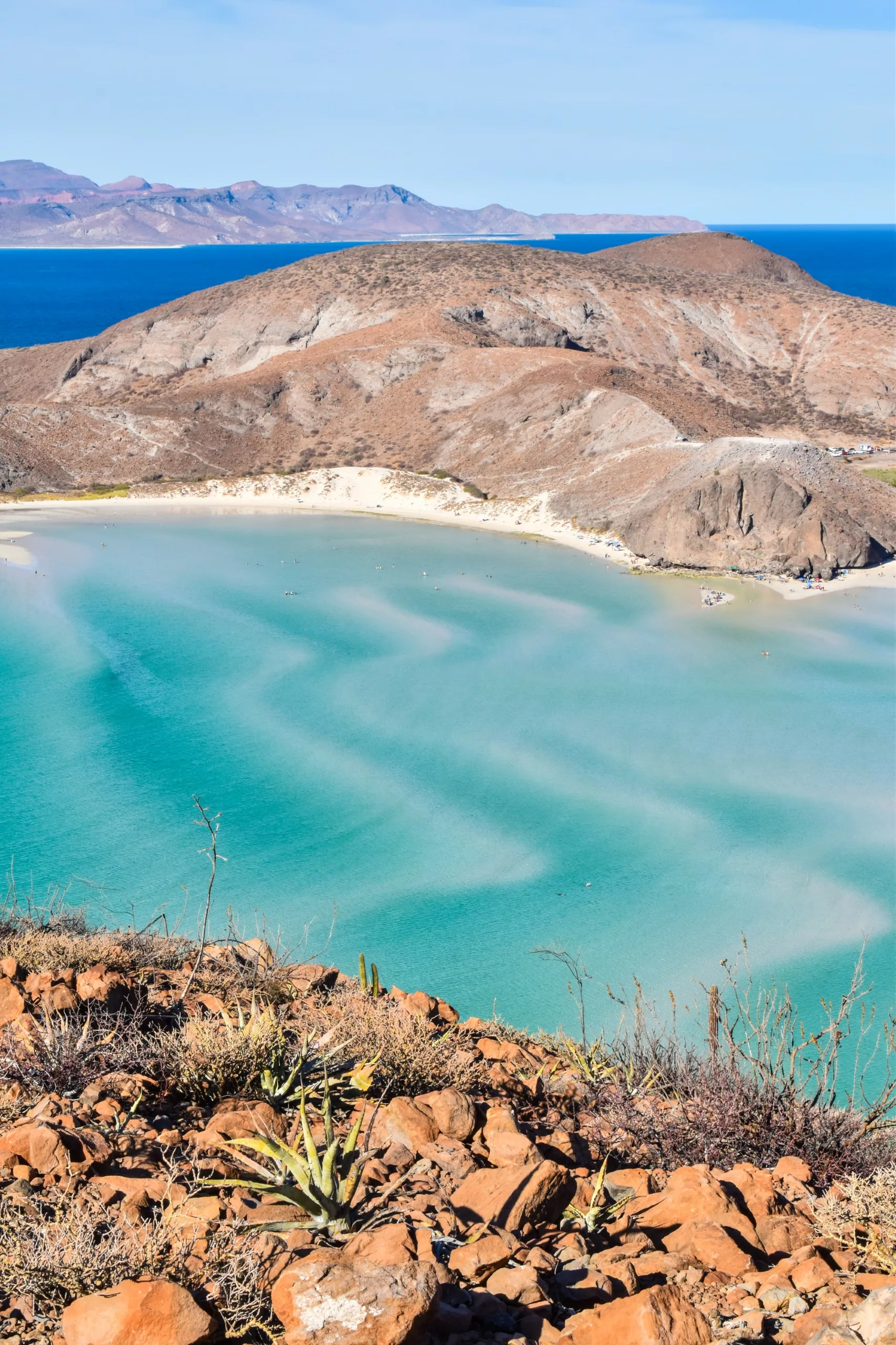 Vue d'une colline rocheuse surplombant la baie turquoise de La Paz, au Mexique, avec de douces vagues, une plage de sable, des montagnes brunes et arides en arrière-plan, et des plantes désertiques au premier plan, sous un ciel bleu clair.