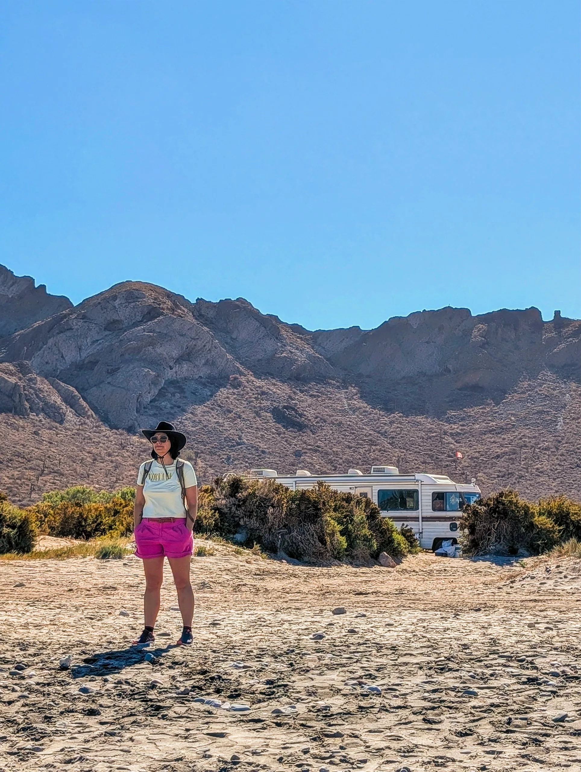 Une personne portant un chapeau, des lunettes de soleil et un short rose se tient sur un terrain sablonneux parsemé d'arbustes. En arrière-plan, un camping-car est garé devant des montagnes rocheuses.