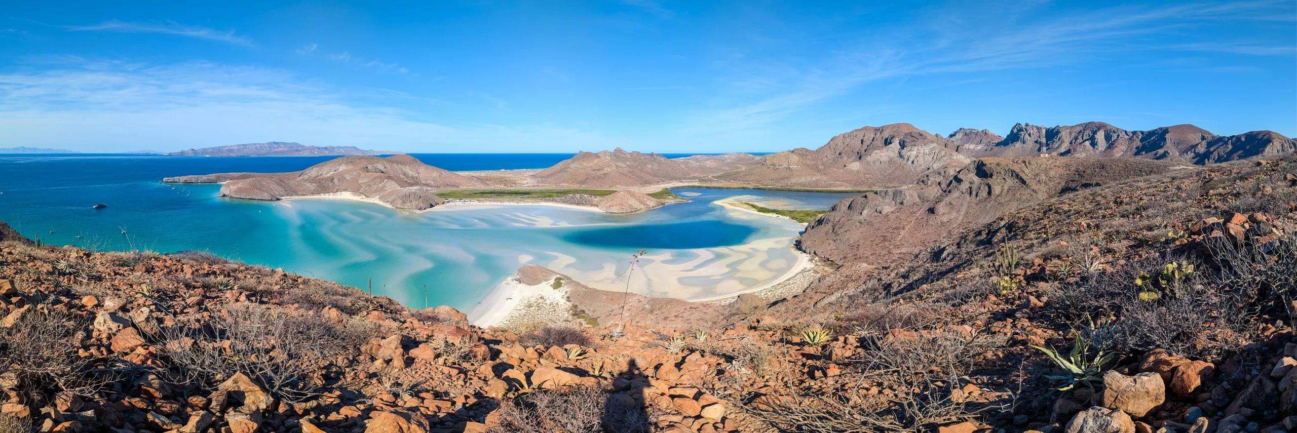 Vue panoramique d'un paysage côtier avec une eau turquoise, des plages de sable et des collines rocheuses sous un ciel bleu clair. De la végétation est visible au premier plan parmi les rochers.